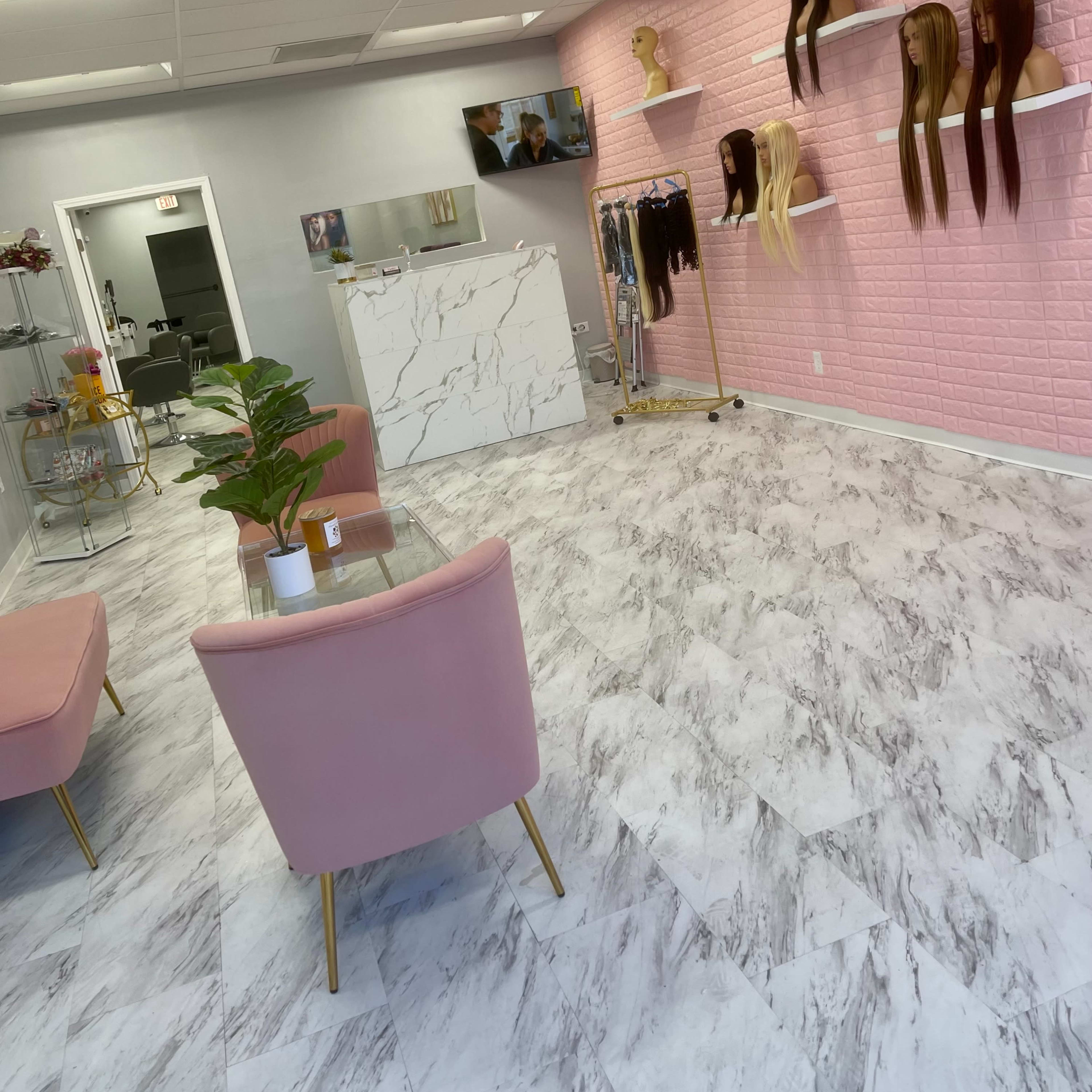 A modern hair salon with a pink accent wall, shelves displaying various wigs, a seating area with pink chairs, and a decorative plant.