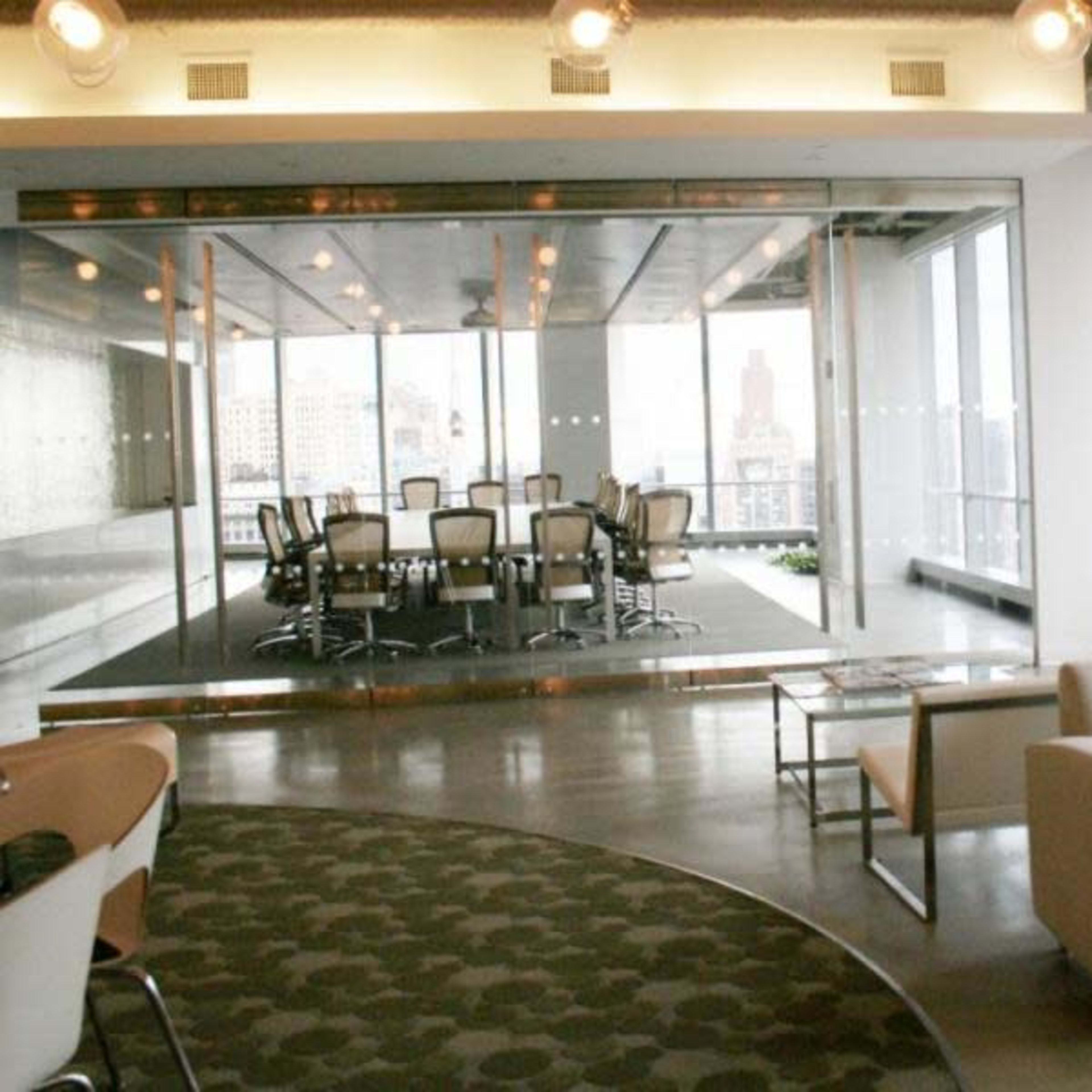 A modern office space features a glass-walled conference room with a large table and chairs, adjacent to a seating area with a couch.