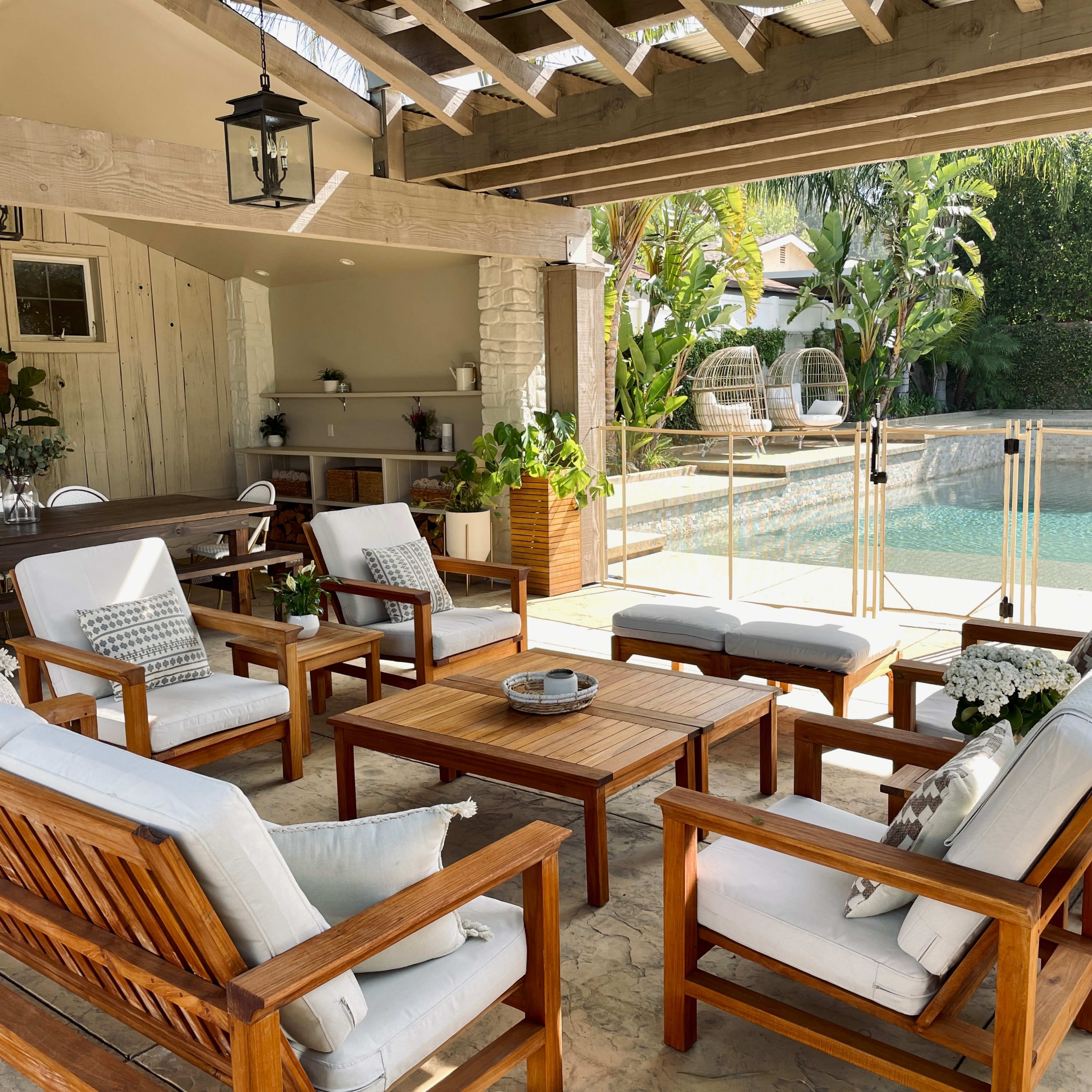 A spacious outdoor seating area features wooden furniture arranged around a central table, overlooking a pool surrounded by greenery.