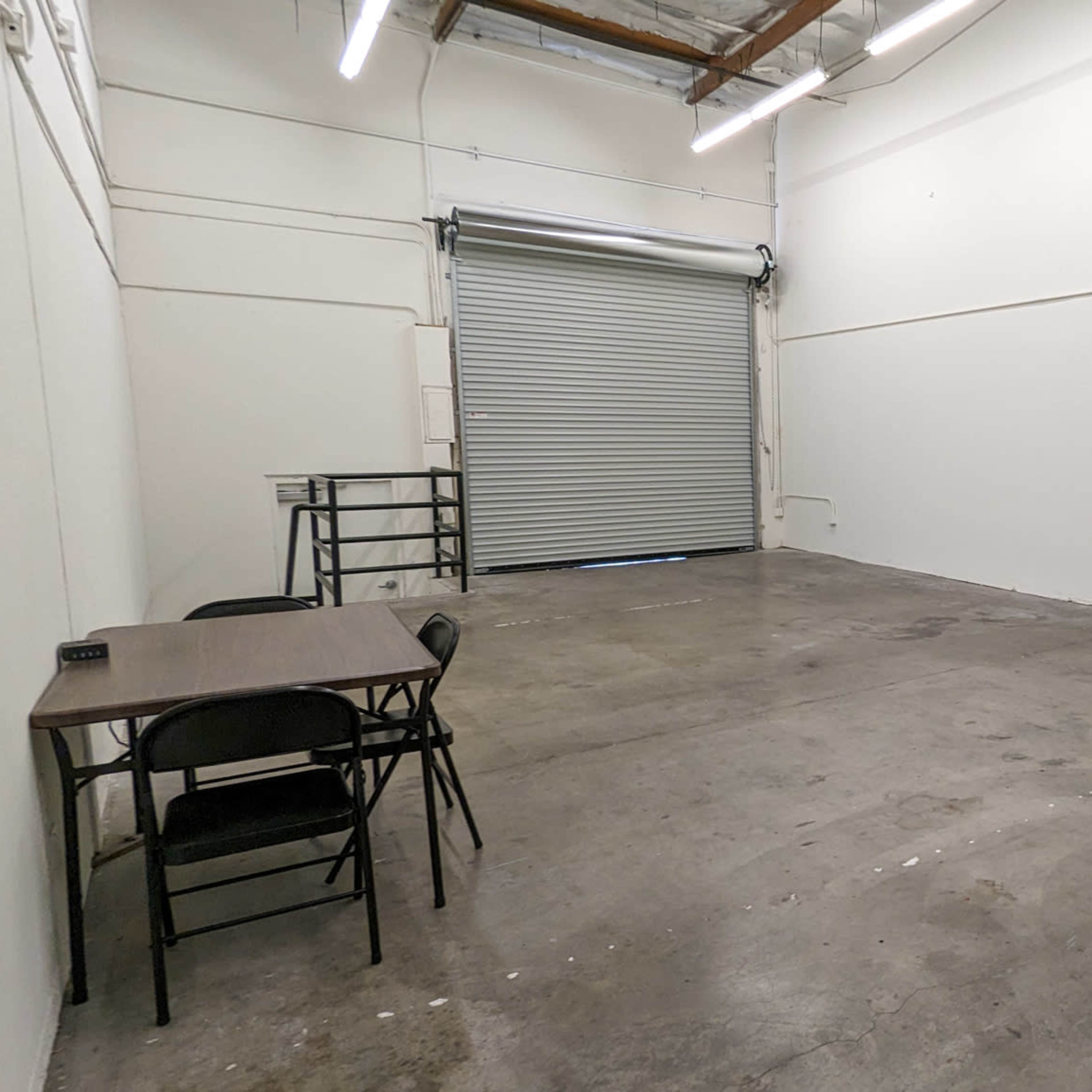 A sparsely furnished room with a concrete floor, a table and chairs, a closed garage door, and several standing light fixture stands.