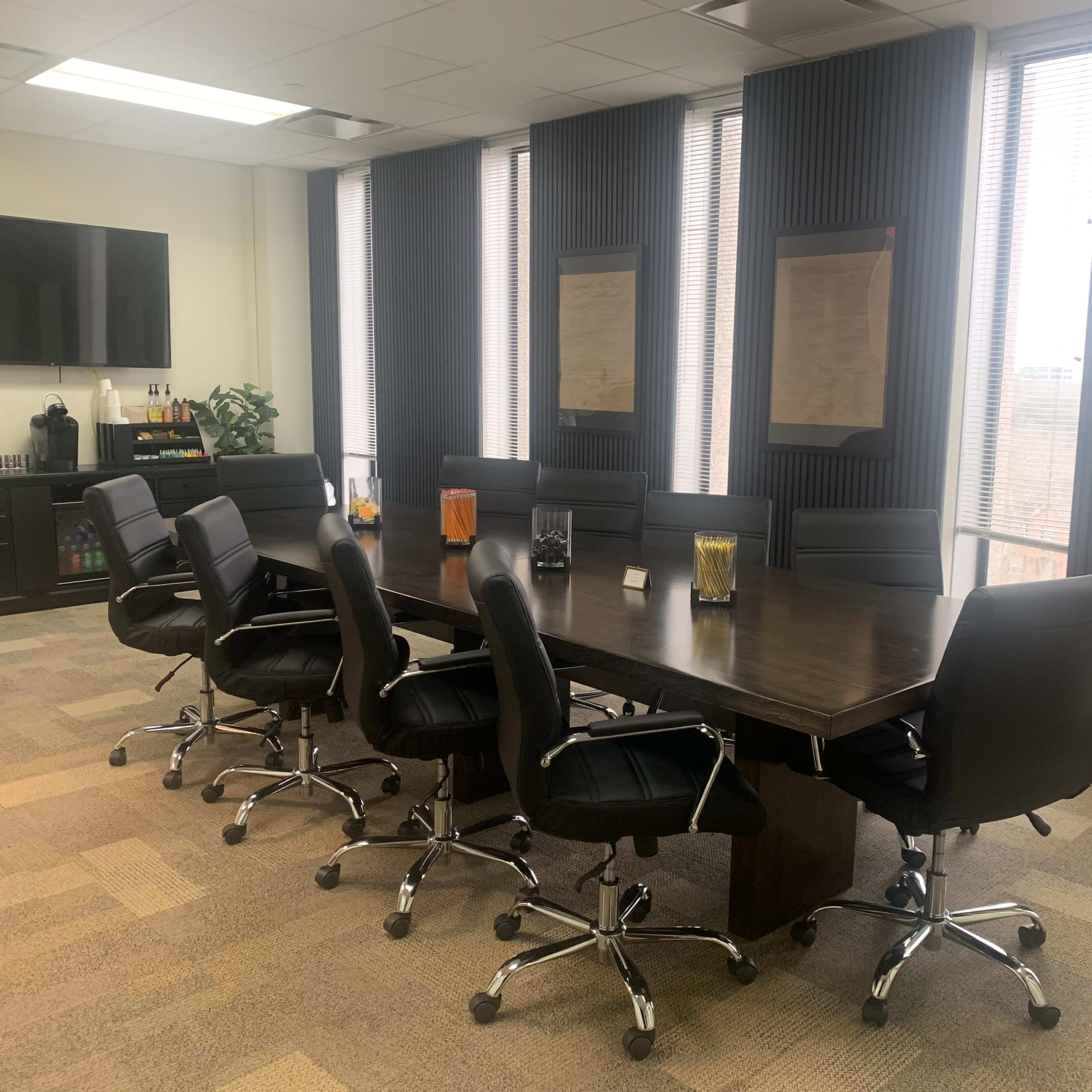 A modern conference room with a large dark wooden table surrounded by black leather chairs, and large windows displaying a city view.