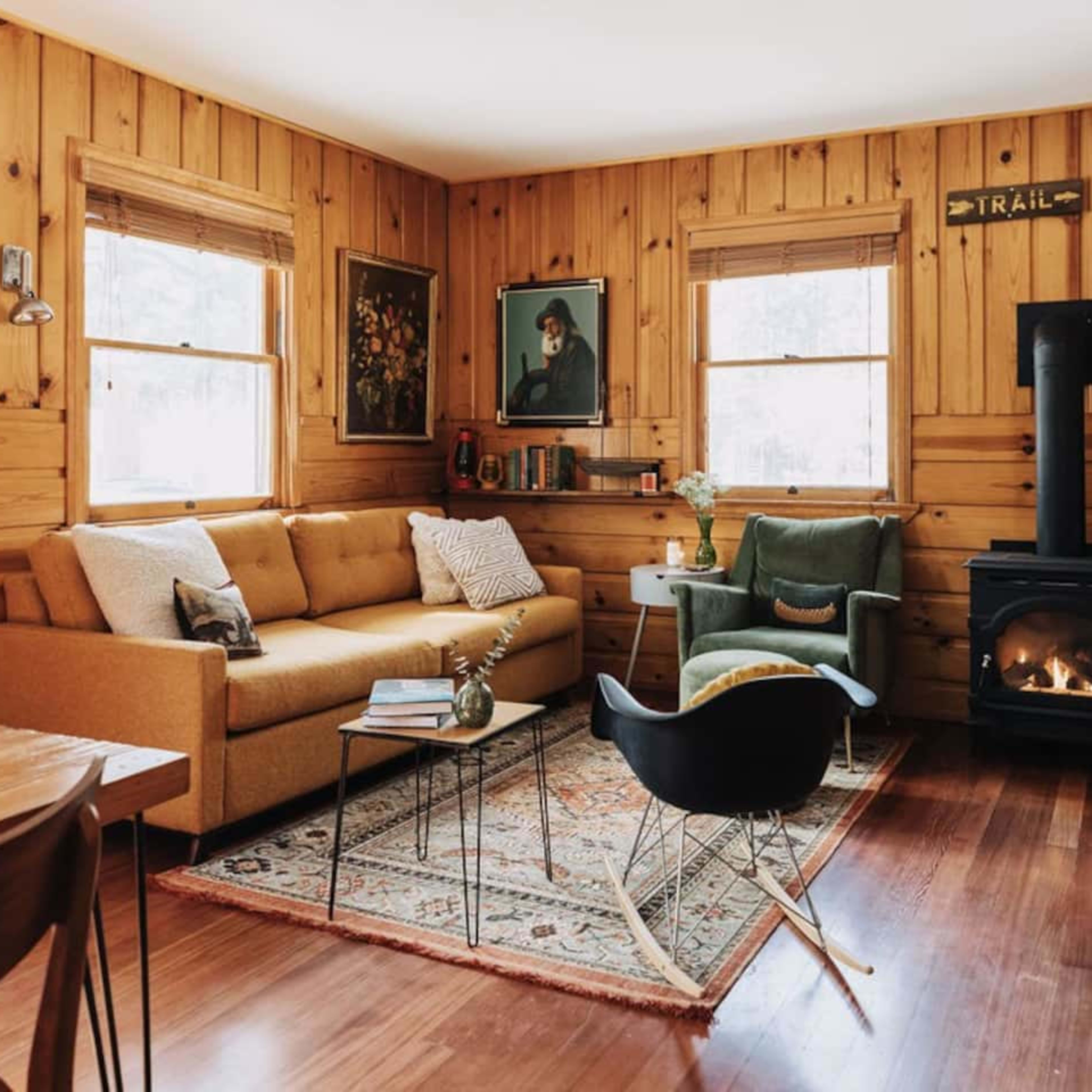 A cozy living room with wooden walls, a sofa, an armchair, a coffee table, and a small stove by the window.