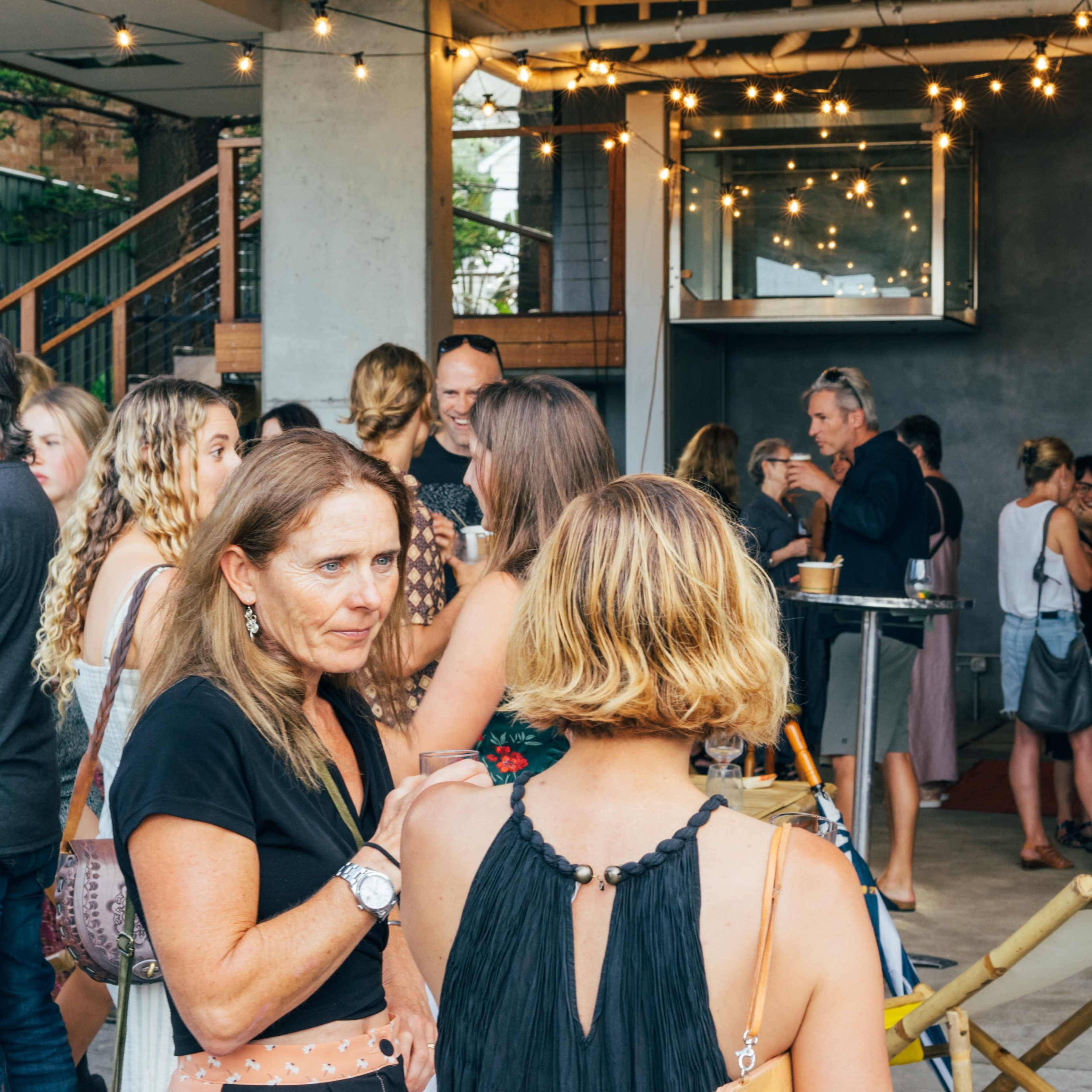 A group of people gathers in a well-lit space, engaging in conversation while surrounded by string lights and a bar area.