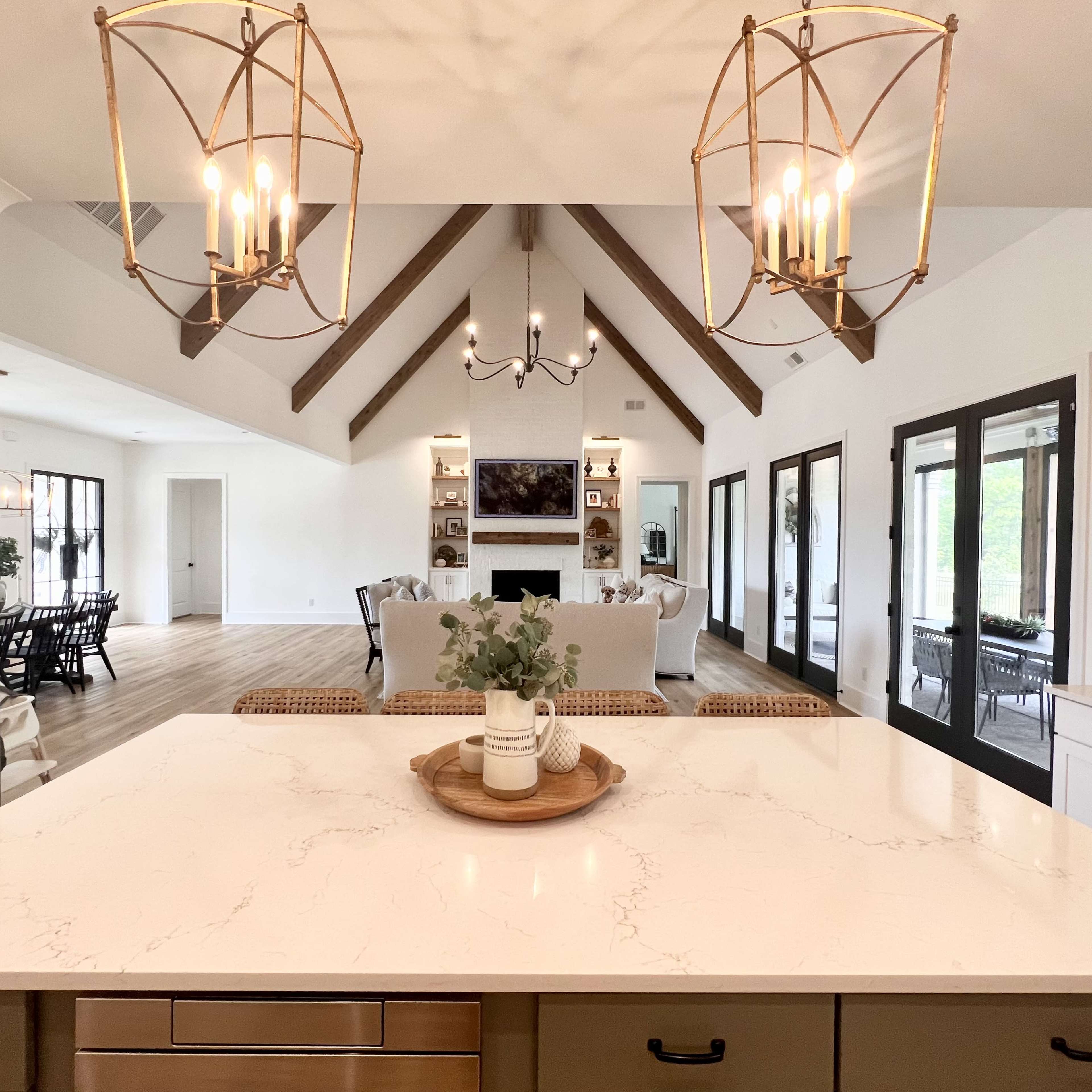 A modern kitchen features a marble island and pendant lights, leading into a spacious living area with a high, beamed ceiling and large windows.