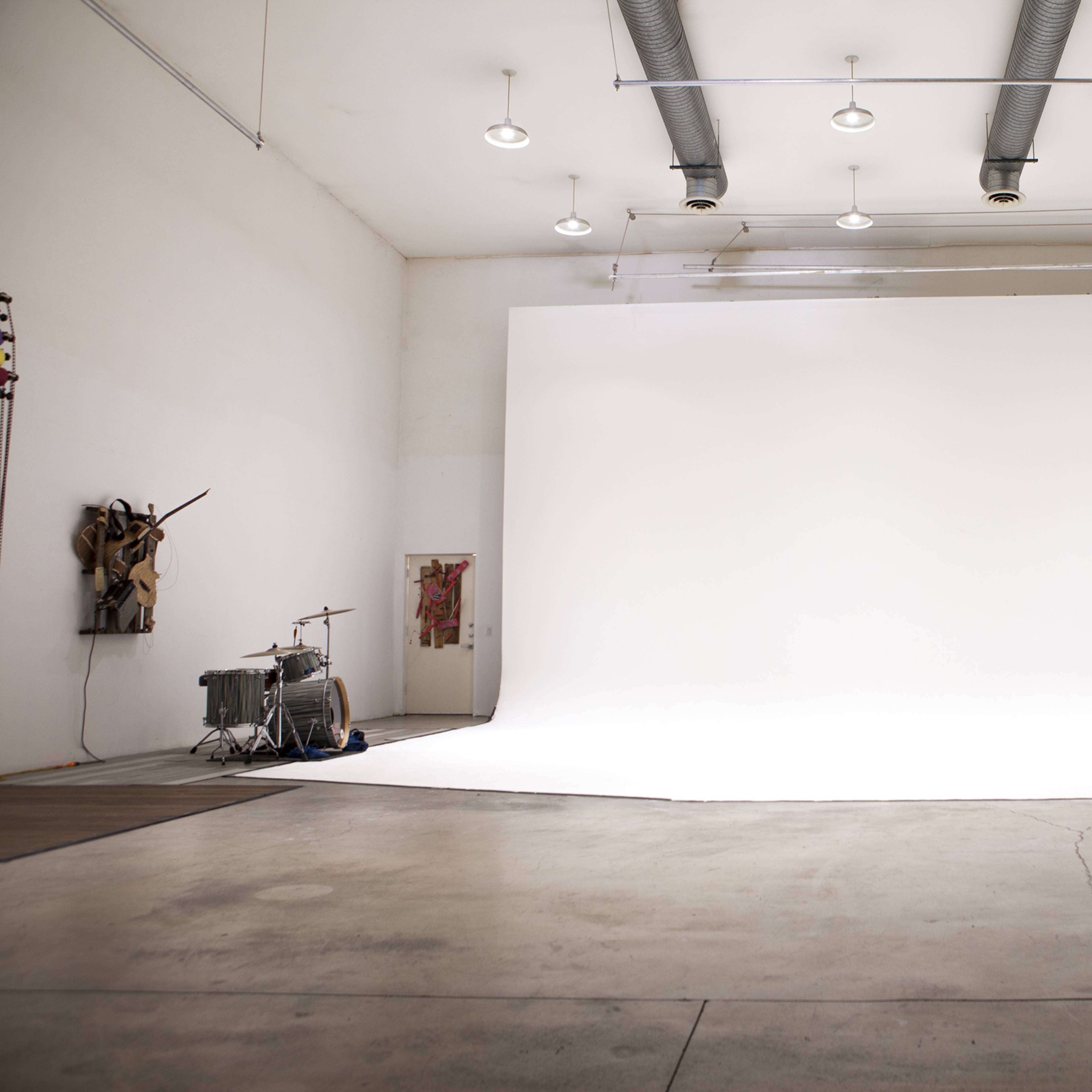 A large, empty studio space with white walls and a seamless white backdrop, featuring industrial lighting and a drum set in the corner.