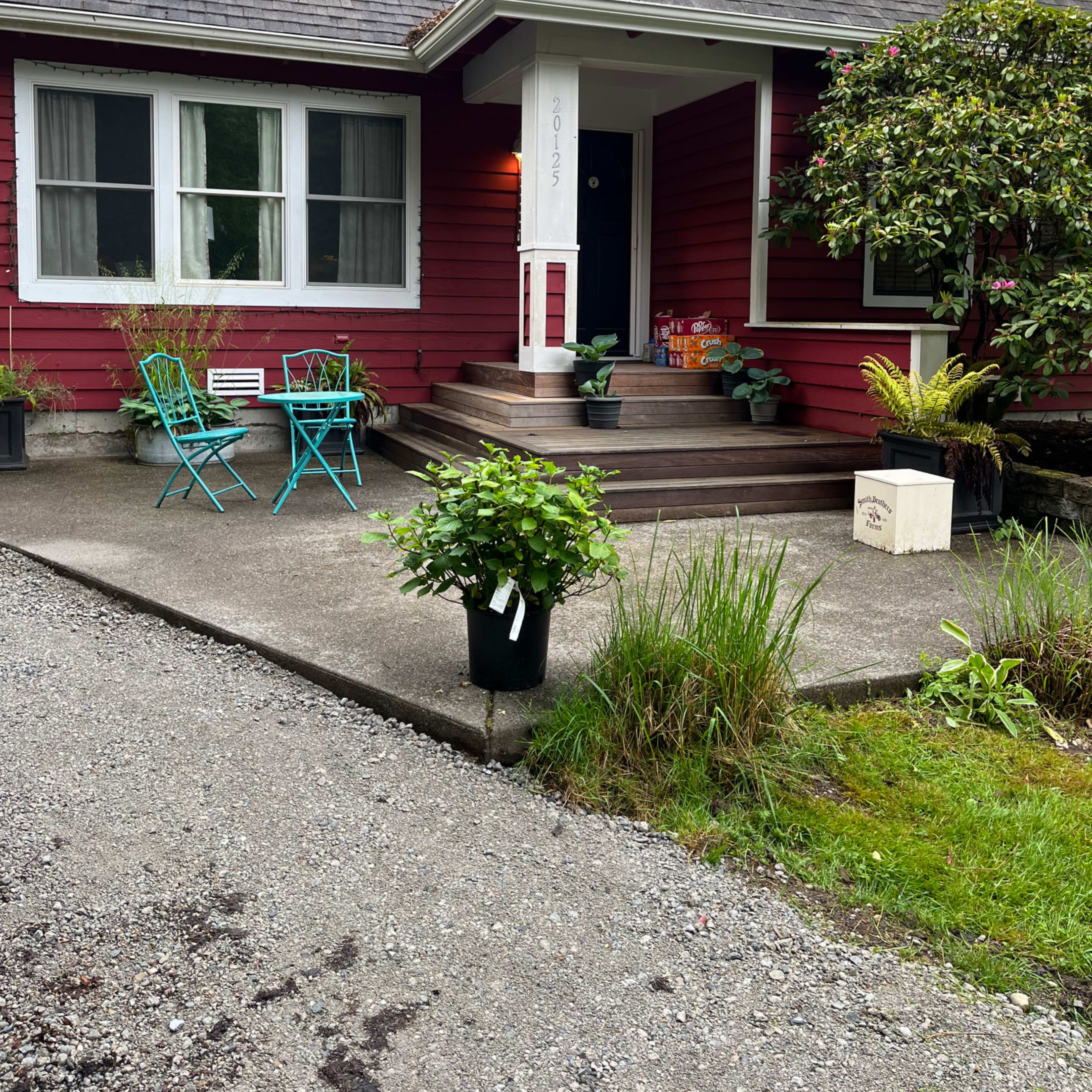 A red house with a front porch features two teal chairs, potted plants, and a gravel driveway lined with greenery.