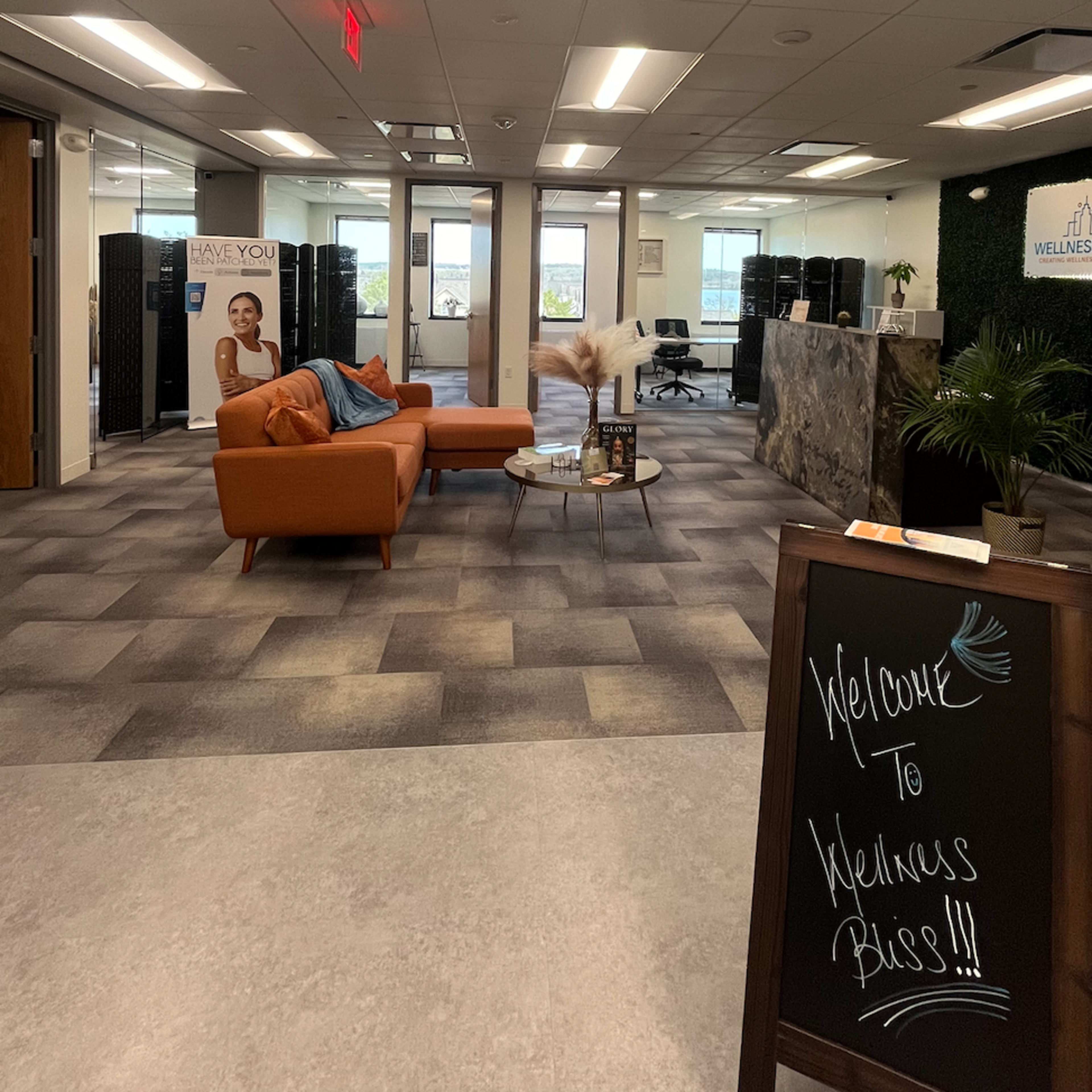 A modern office lobby with an orange couch, a reception desk, and a welcome sign, featuring large windows that provide a view of the outside.