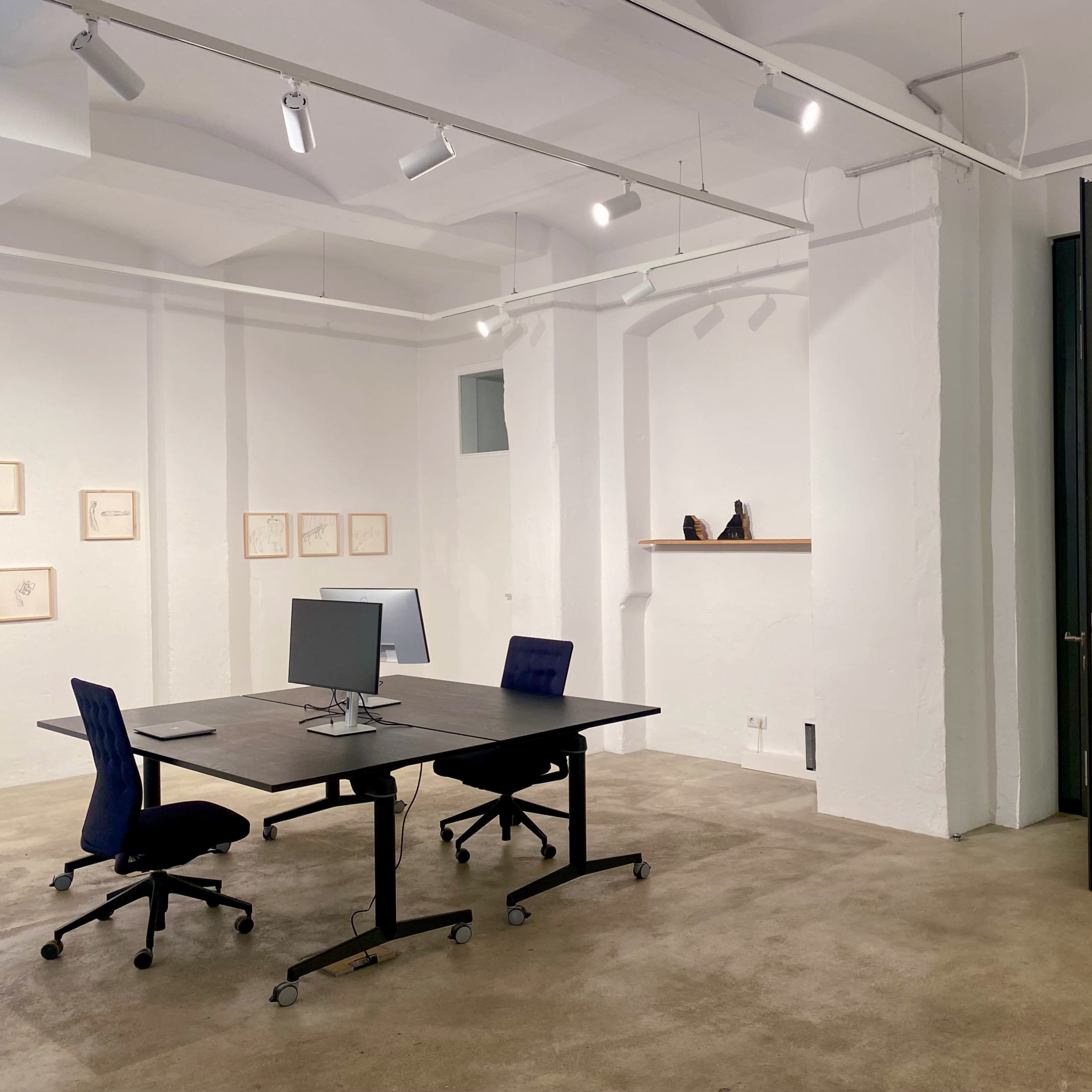 A minimalist office space features a dark table with a computer and two blue chairs, surrounded by white walls and minimal artwork.