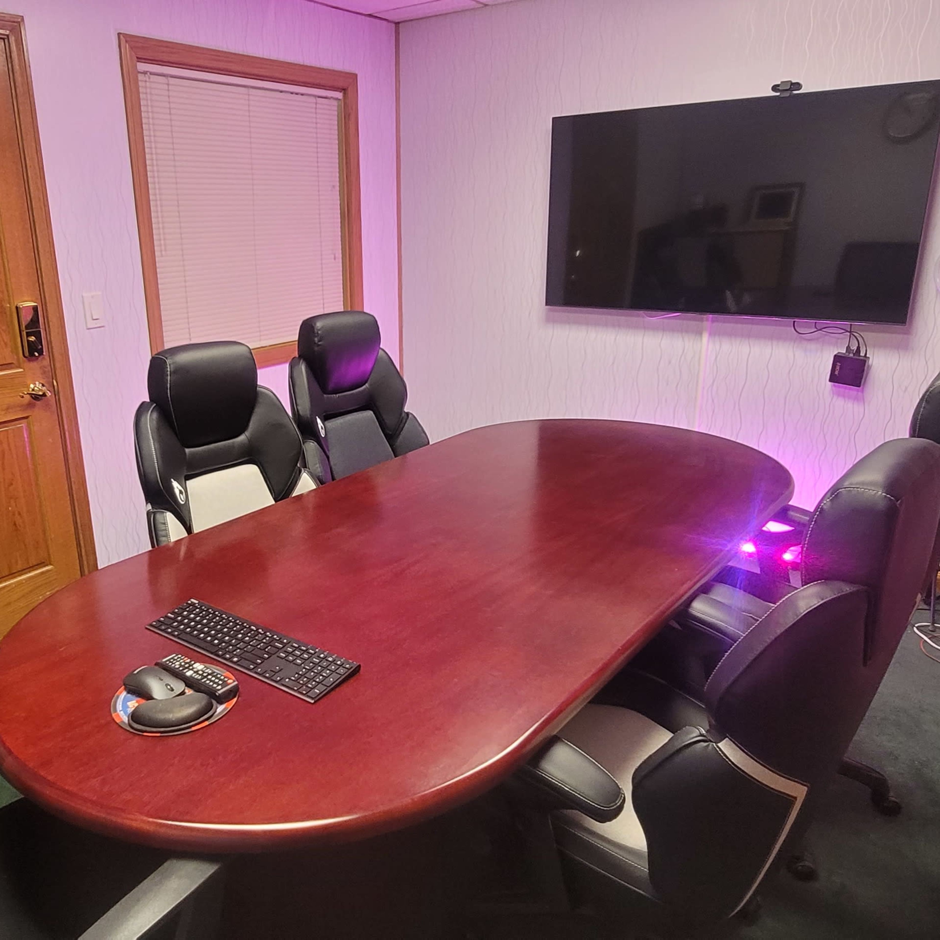 A conference room with a large wooden table surrounded by black chairs, a wall-mounted television, and soft lighting.
