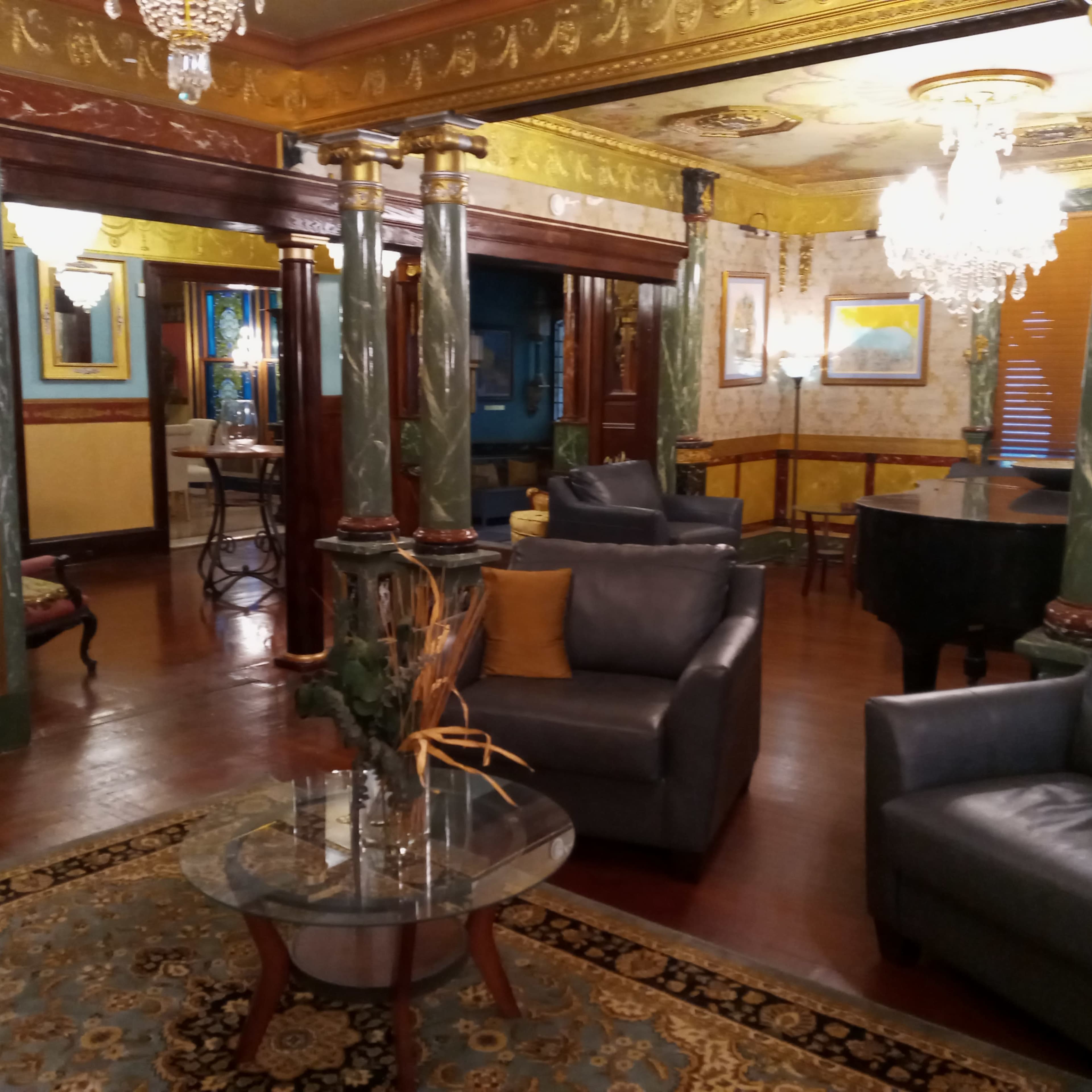A partially furnished room featuring green marble columns, leather sofas, a glass coffee table, and a grand piano in an elegantly decorated setting.
