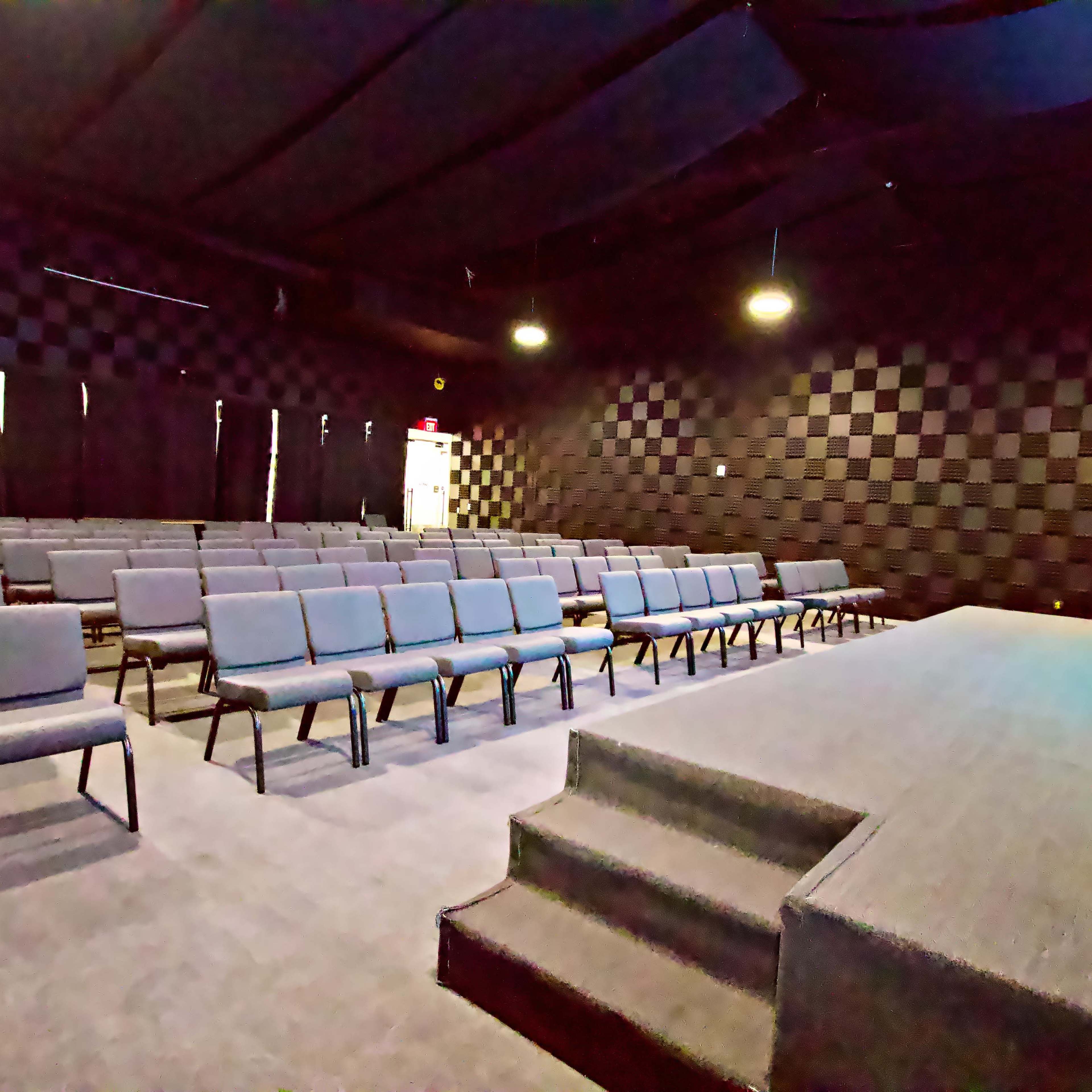 An empty auditorium with rows of gray chairs facing a small stage, all set against a black and gray checkered wall.