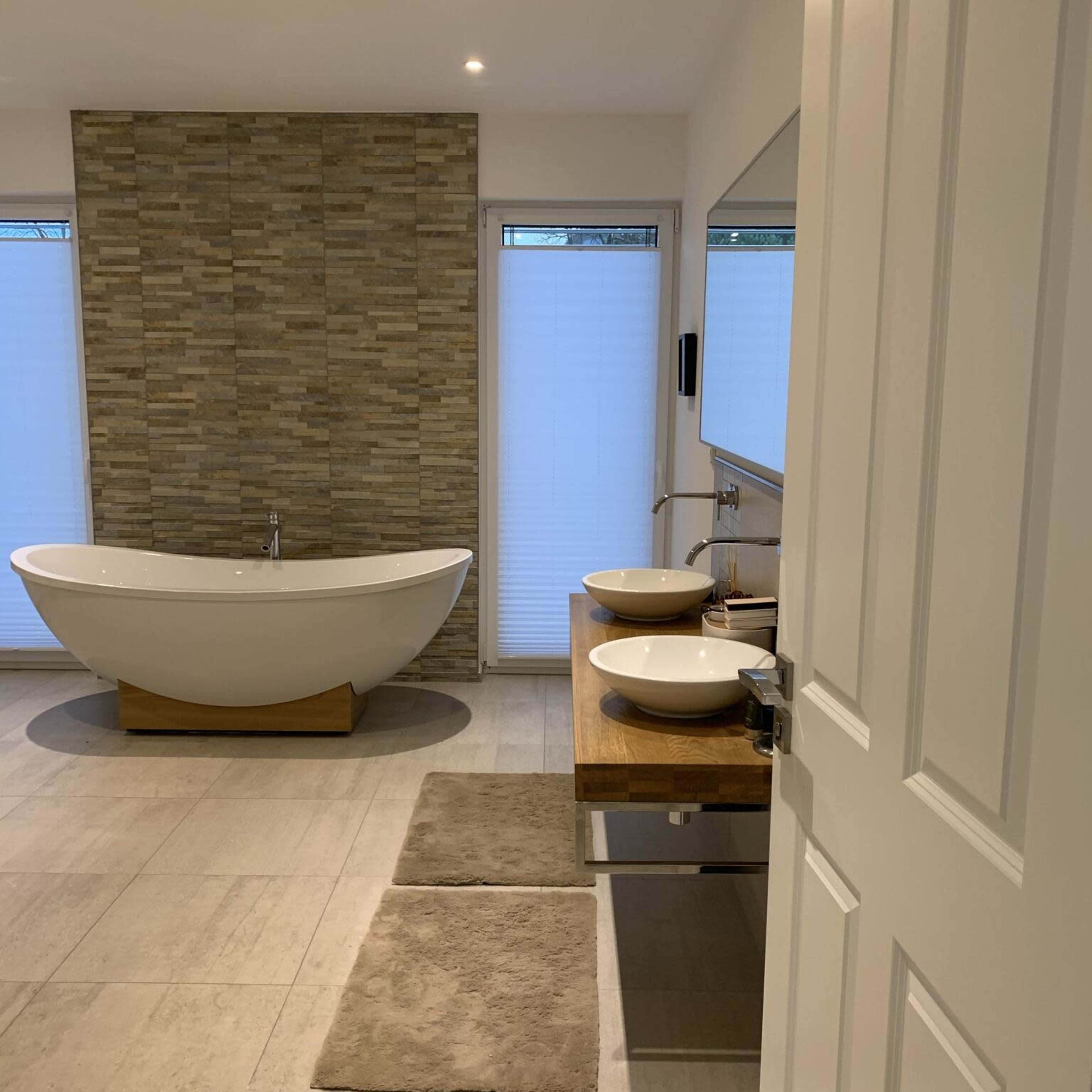 A modern bathroom featuring a freestanding bathtub, two sinks on a wooden countertop, and a stone wall backdrop.
