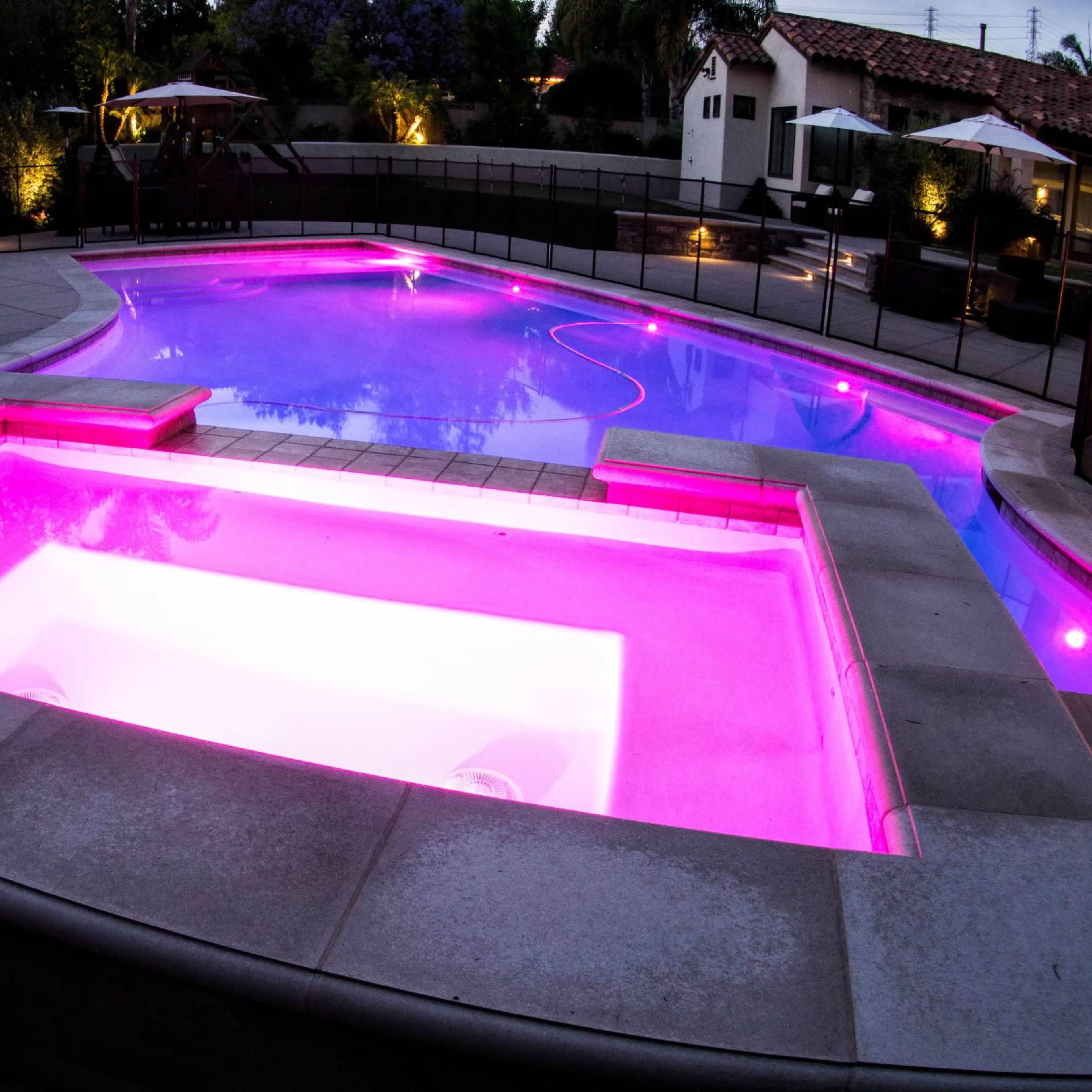 A swimming pool with a pink-lit spa area, surrounded by landscaped gardens and lounge chairs in a residential backyard.