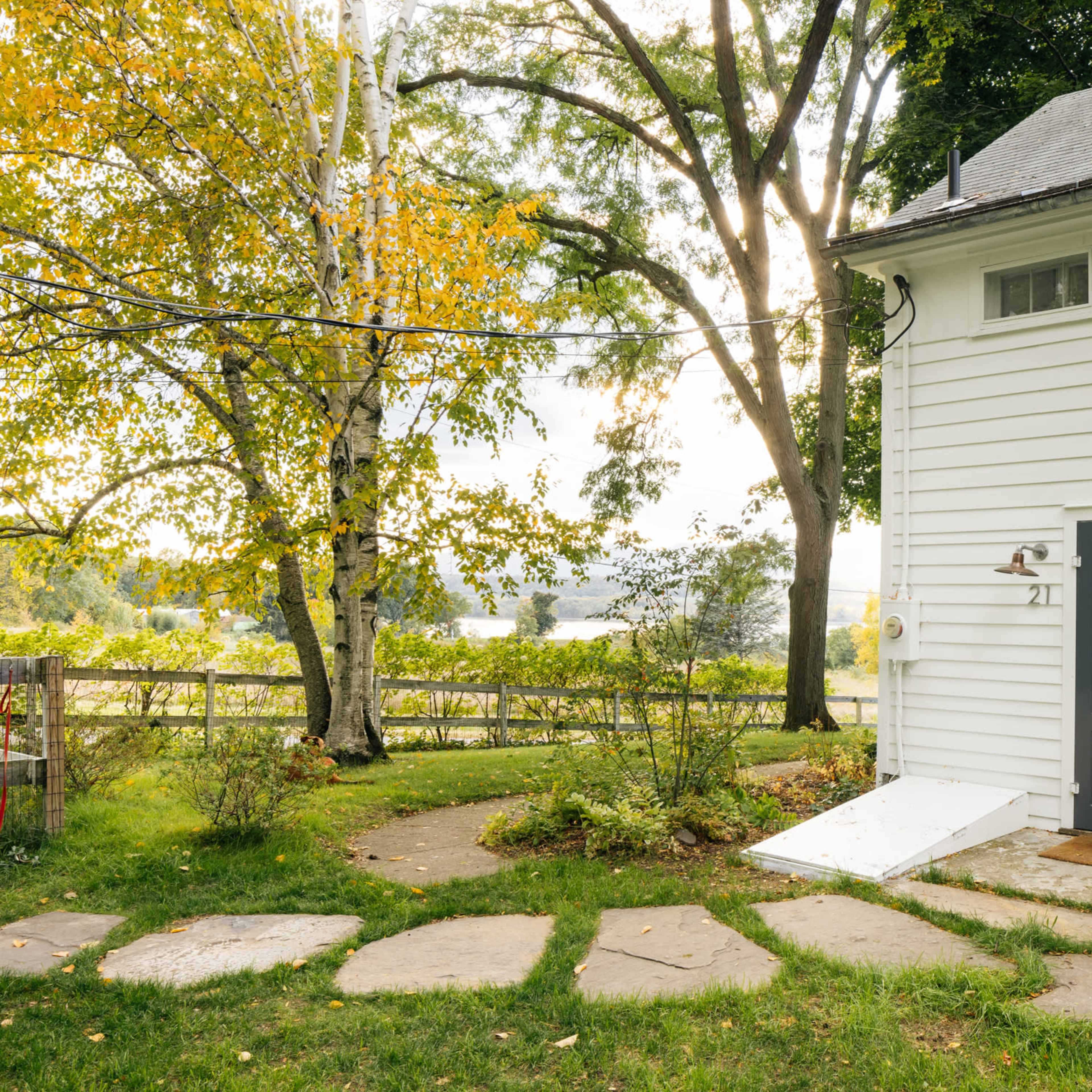 A gray house with white siding has a main entrance door and a stone pathway leading to it, surrounded by trees and a grassy area.