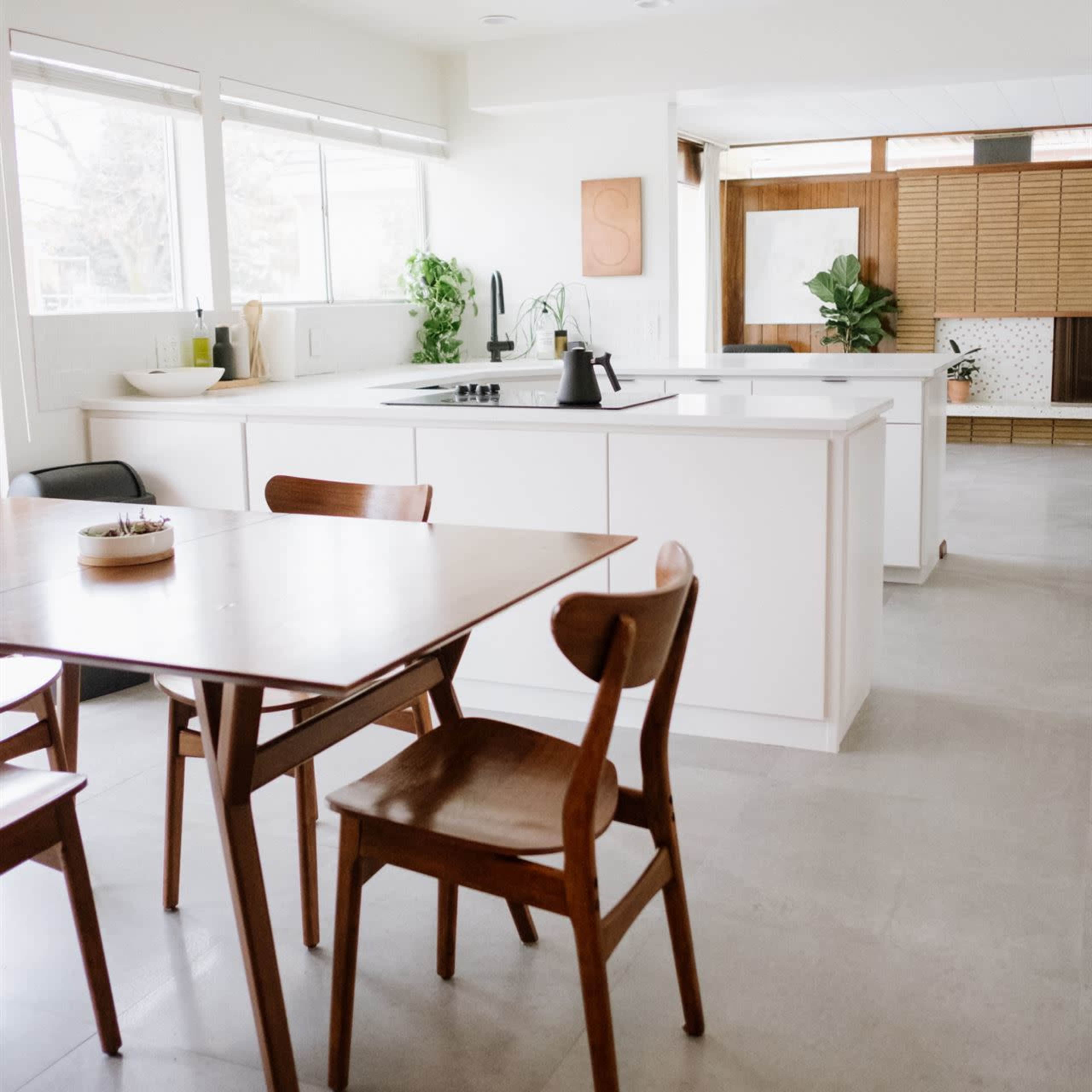 A modern kitchen features a wooden dining table with chairs, a sink, and plants by the windows, with light-colored cabinets and a spacious layout.