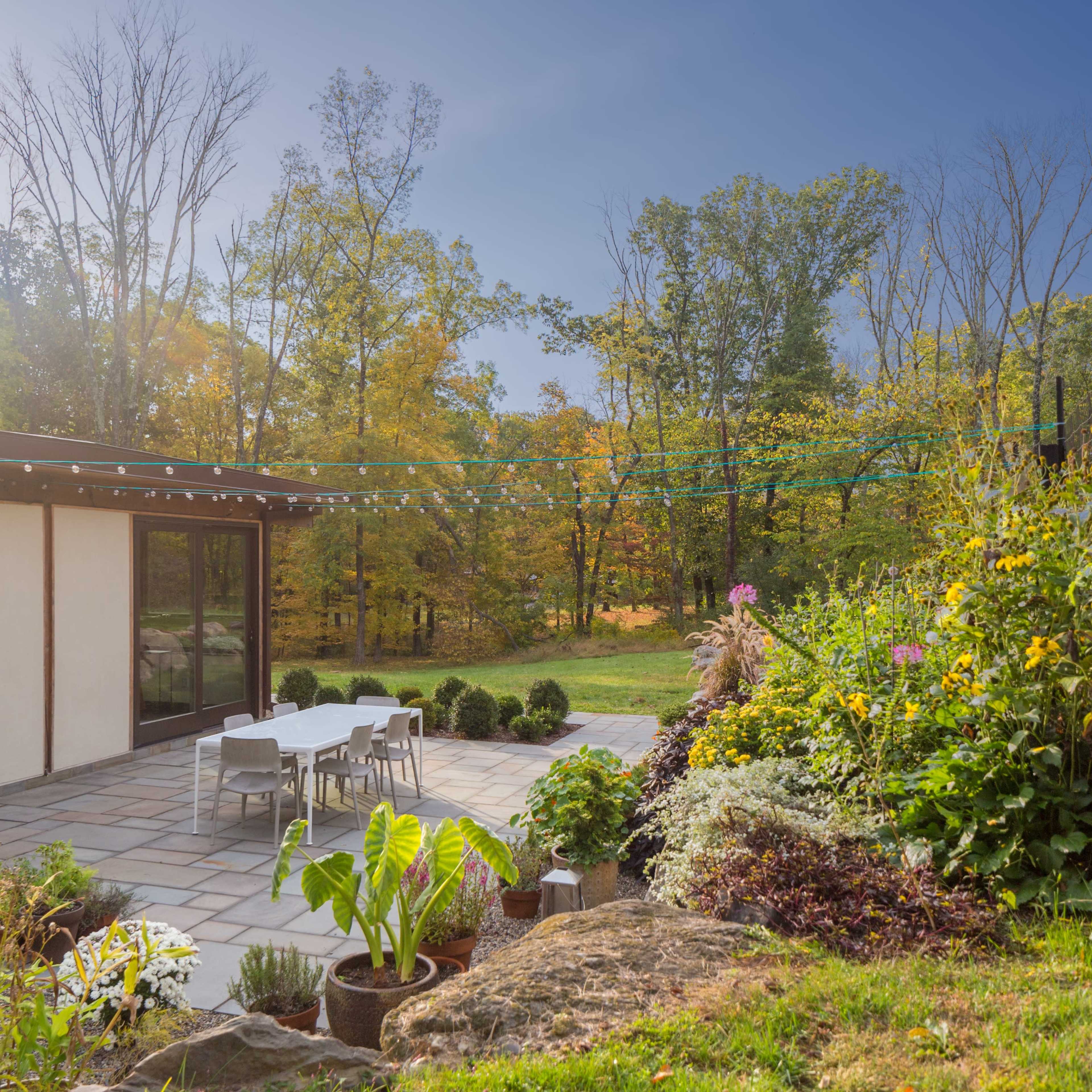 A modern home features a stone patio with white furniture, surrounded by lush greenery and trees in an autumn landscape.