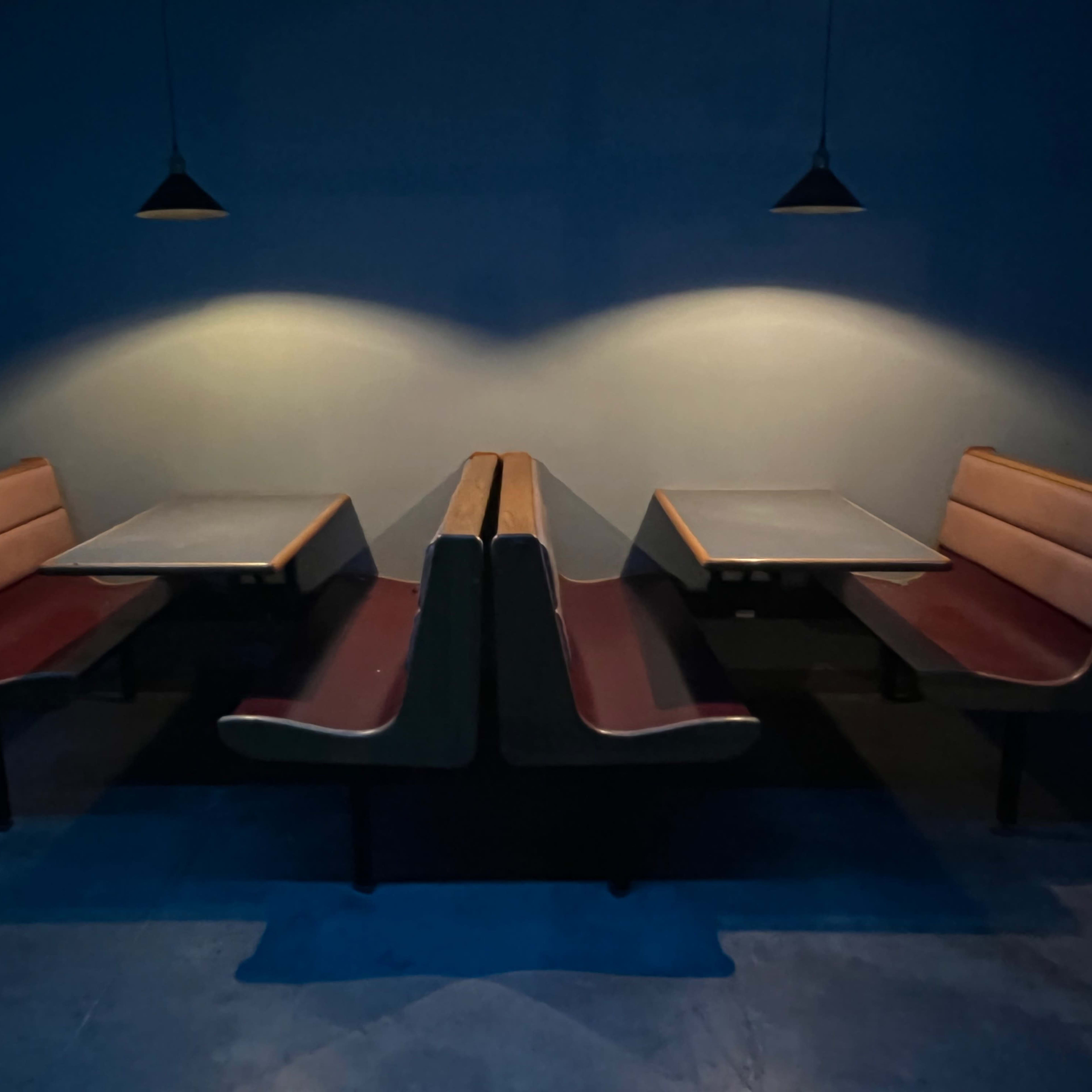 Two booth-style tables with benches are positioned opposite each other in a dimly lit space, illuminated by two pendant lights on the ceiling.
