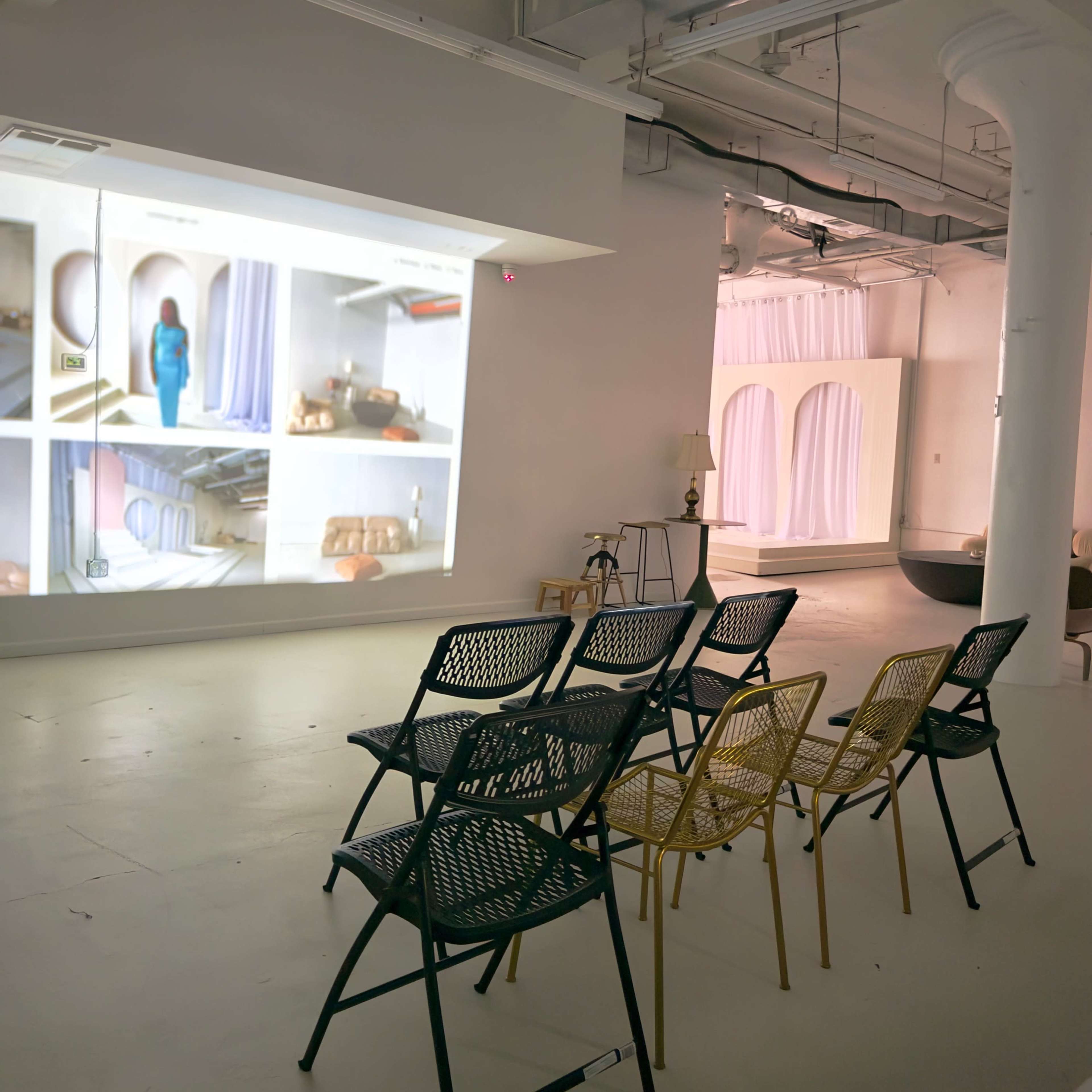 A minimalistic gallery space features a projection displaying images of interiors, with several chairs arranged in front for viewing.