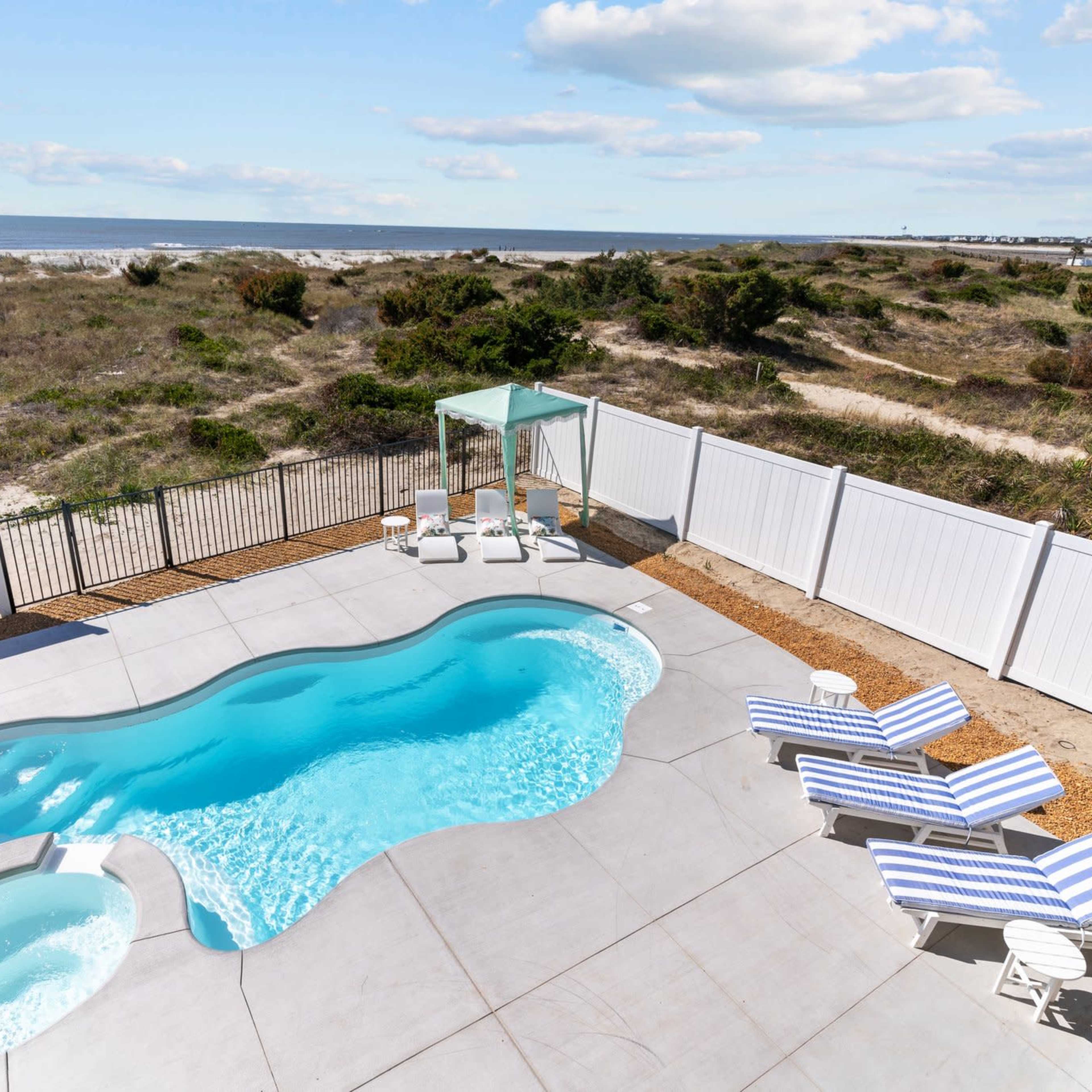 A fenced pool area with lounge chairs, a hot tub, and a view of the beach and greenery in the background.