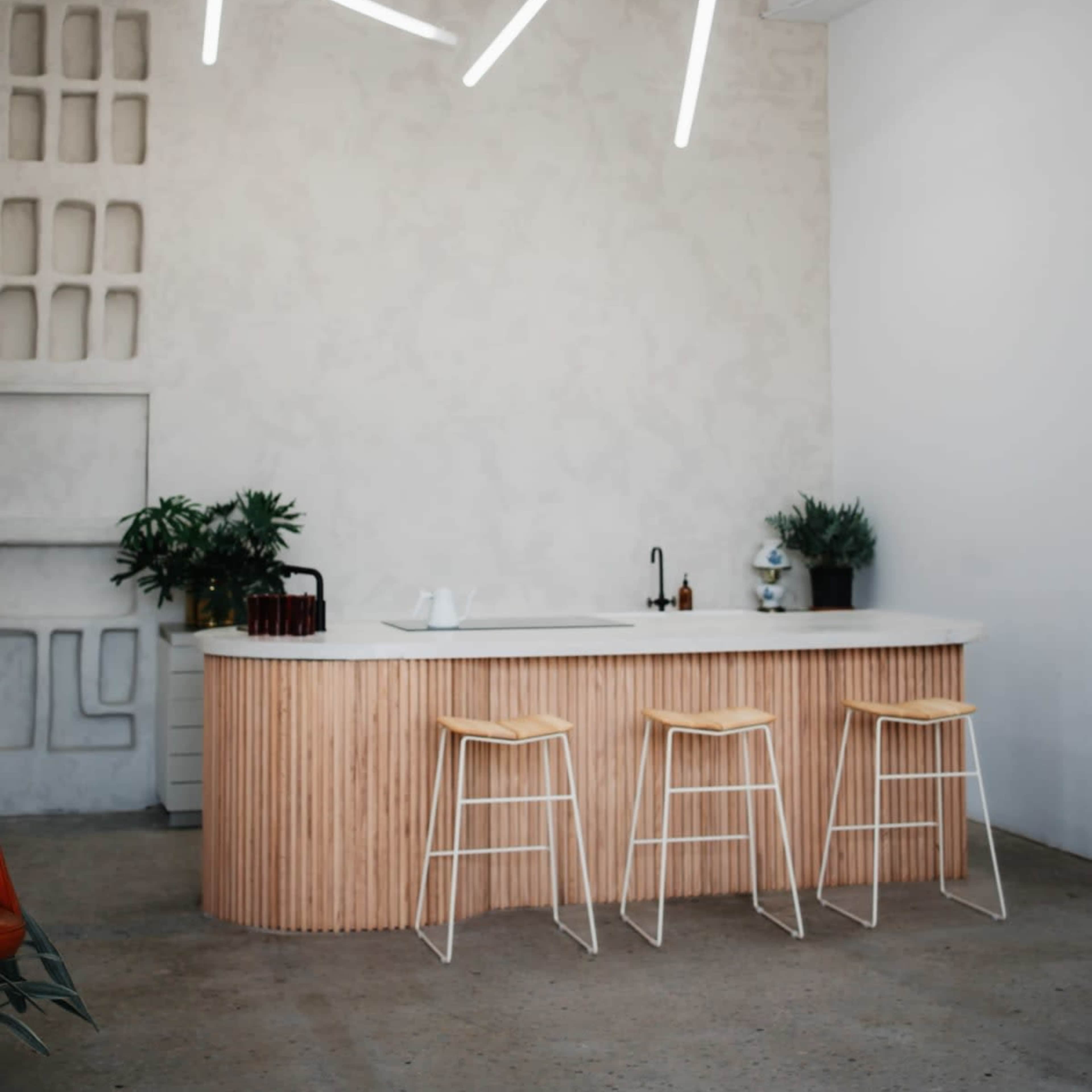 A modern kitchen features a curved wooden bar with three white stools and minimalist lighting above.