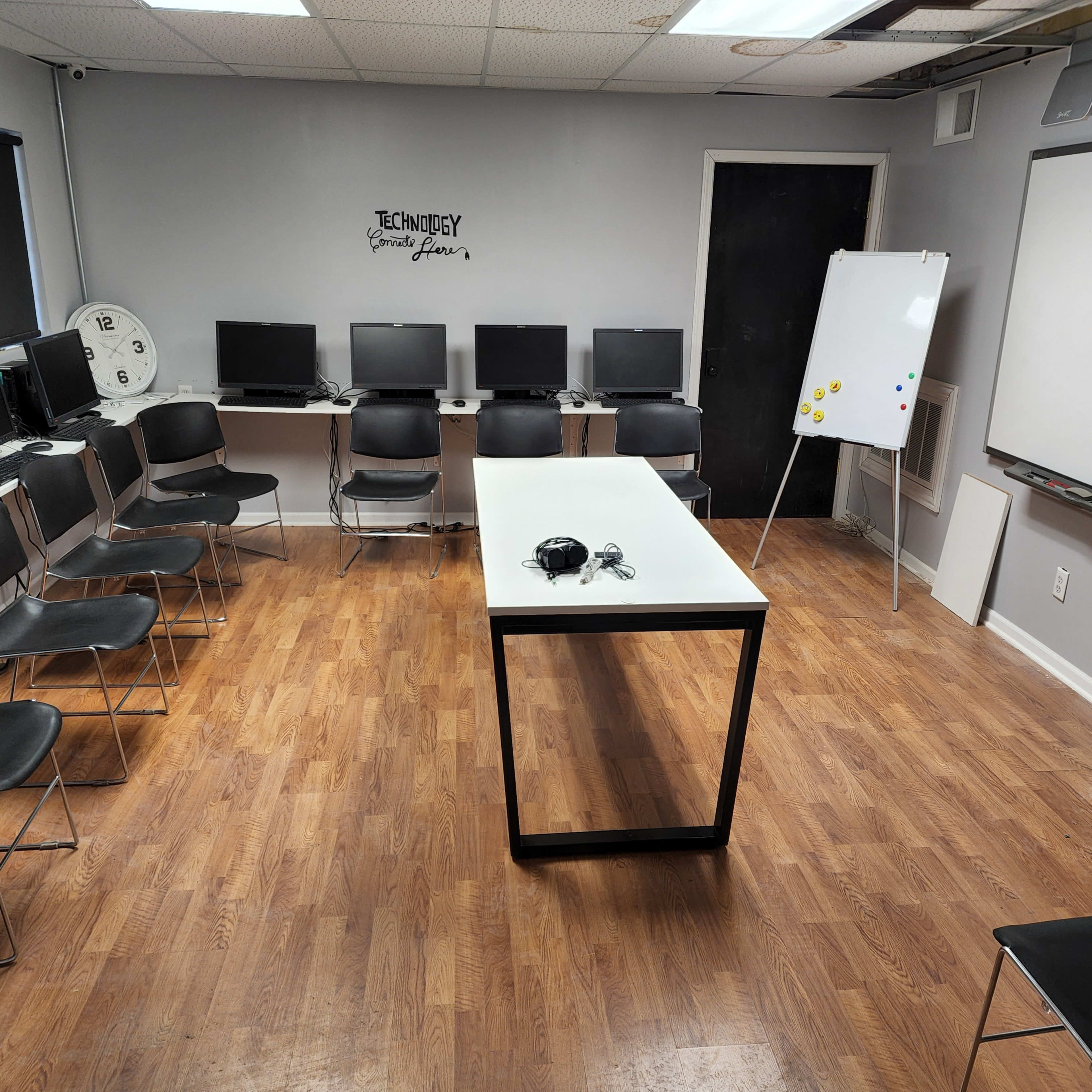 A classroom equipped with several computers and a central table, surrounded by black chairs, along with a whiteboard and a clock on the wall.
