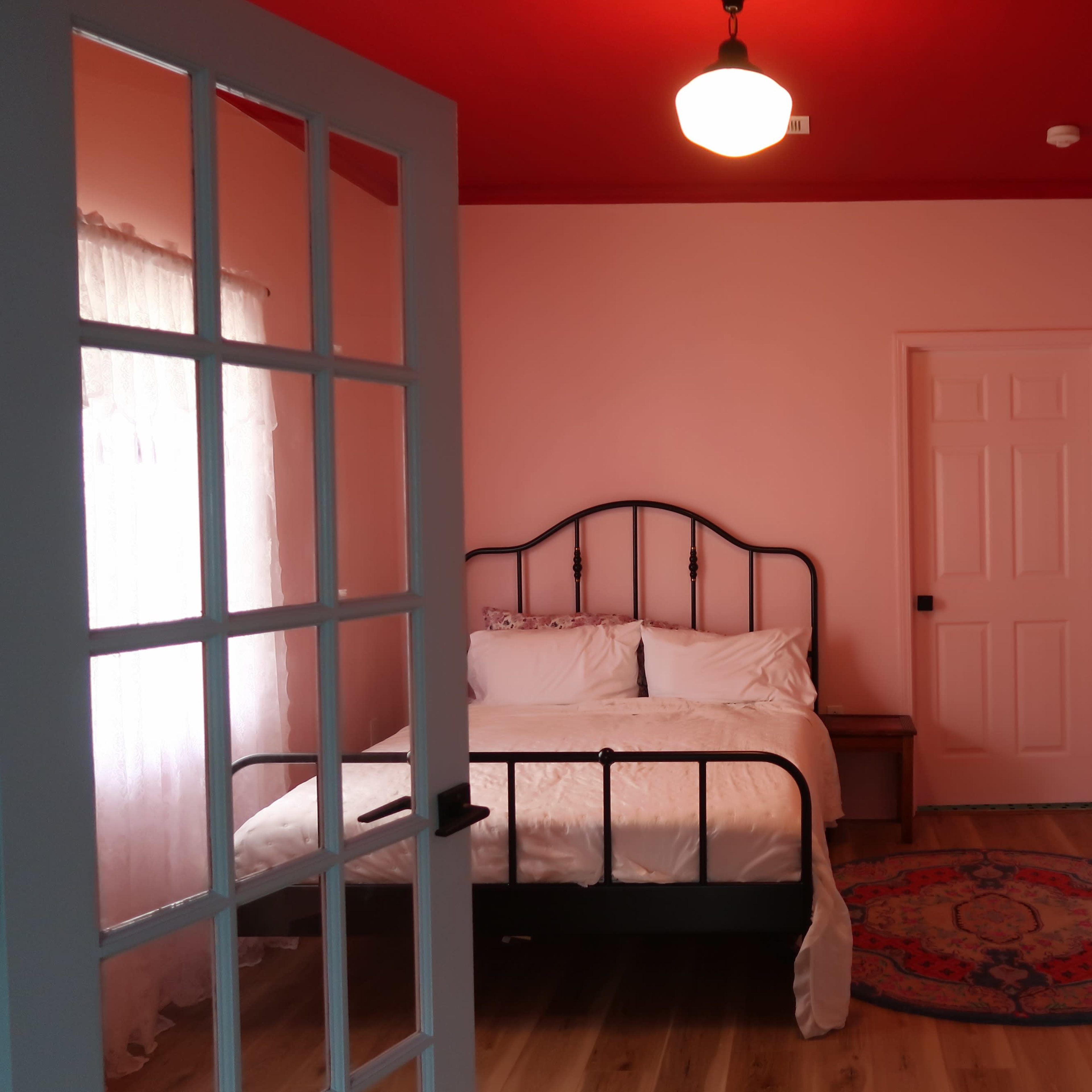A bedroom with a black metal bed frame, white bedding, a red ceiling, and a wooden floor, viewed through an open door.