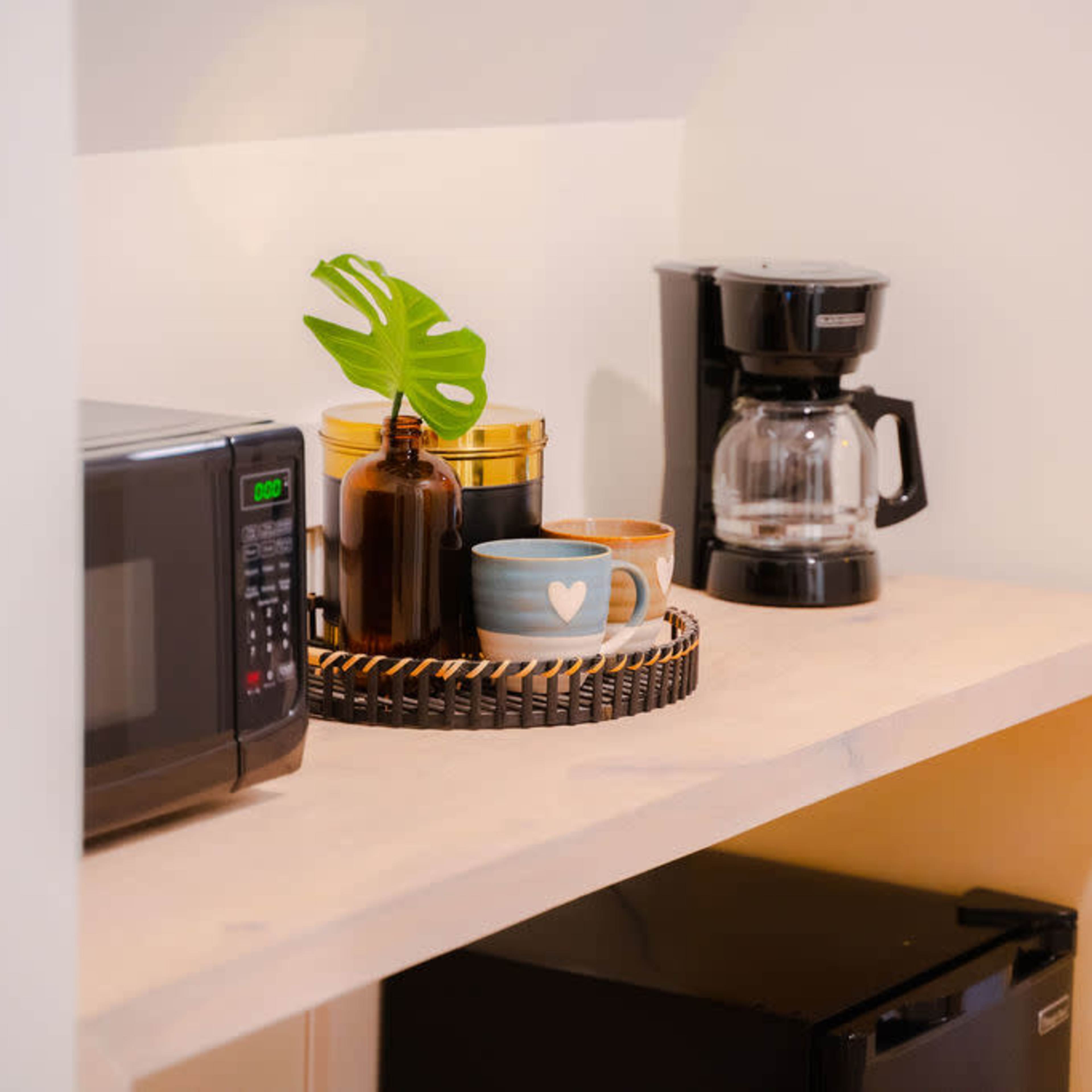 A kitchen countertop displays a microwave, a coffee maker, a small plant, a tray with two mugs, and several jars.
