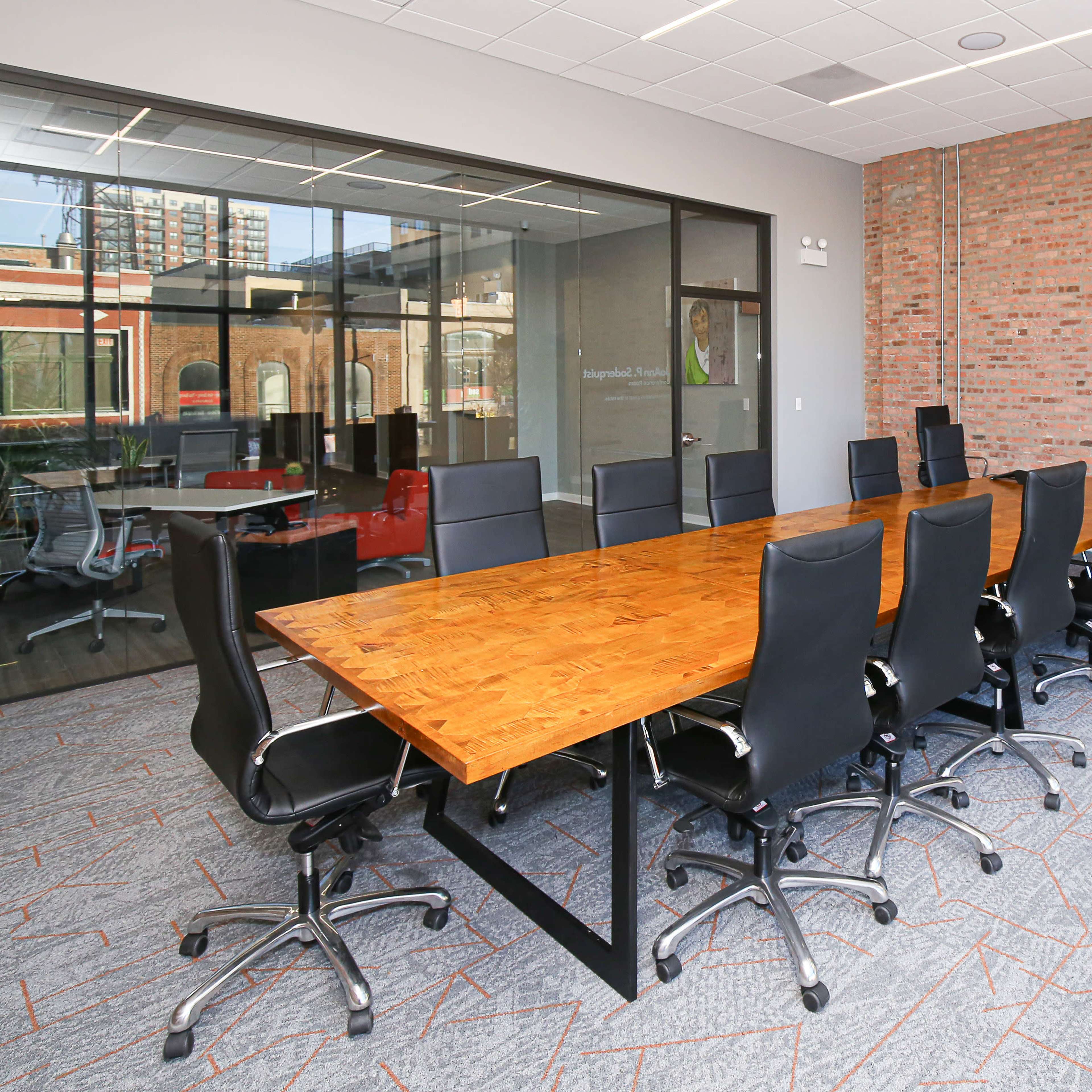 A spacious conference room featuring a long wooden table surrounded by black ergonomic chairs, with a large glass window and a plant in the corner.