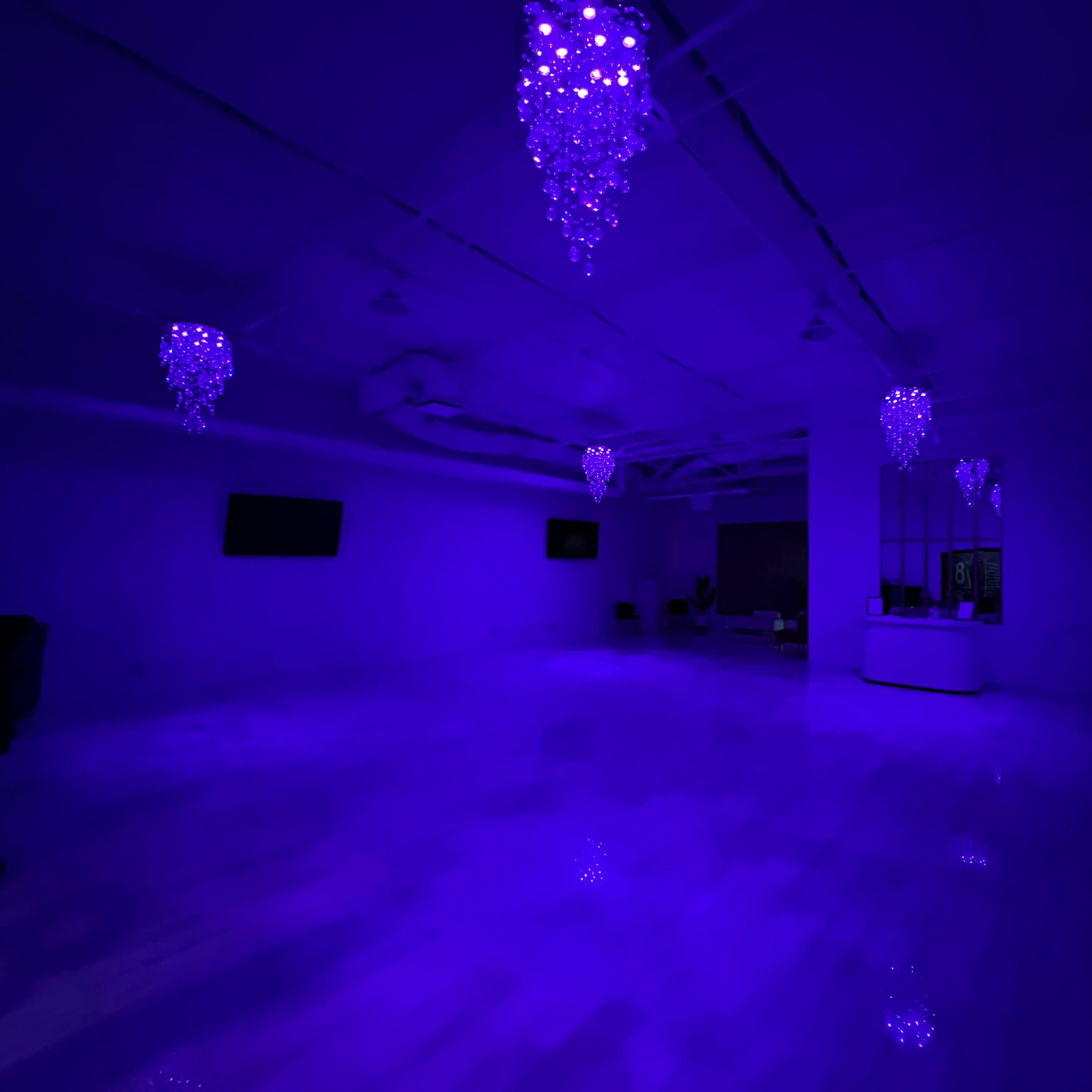 A large, empty room is illuminated in purple light, featuring several chandelier-like fixtures on the ceiling.