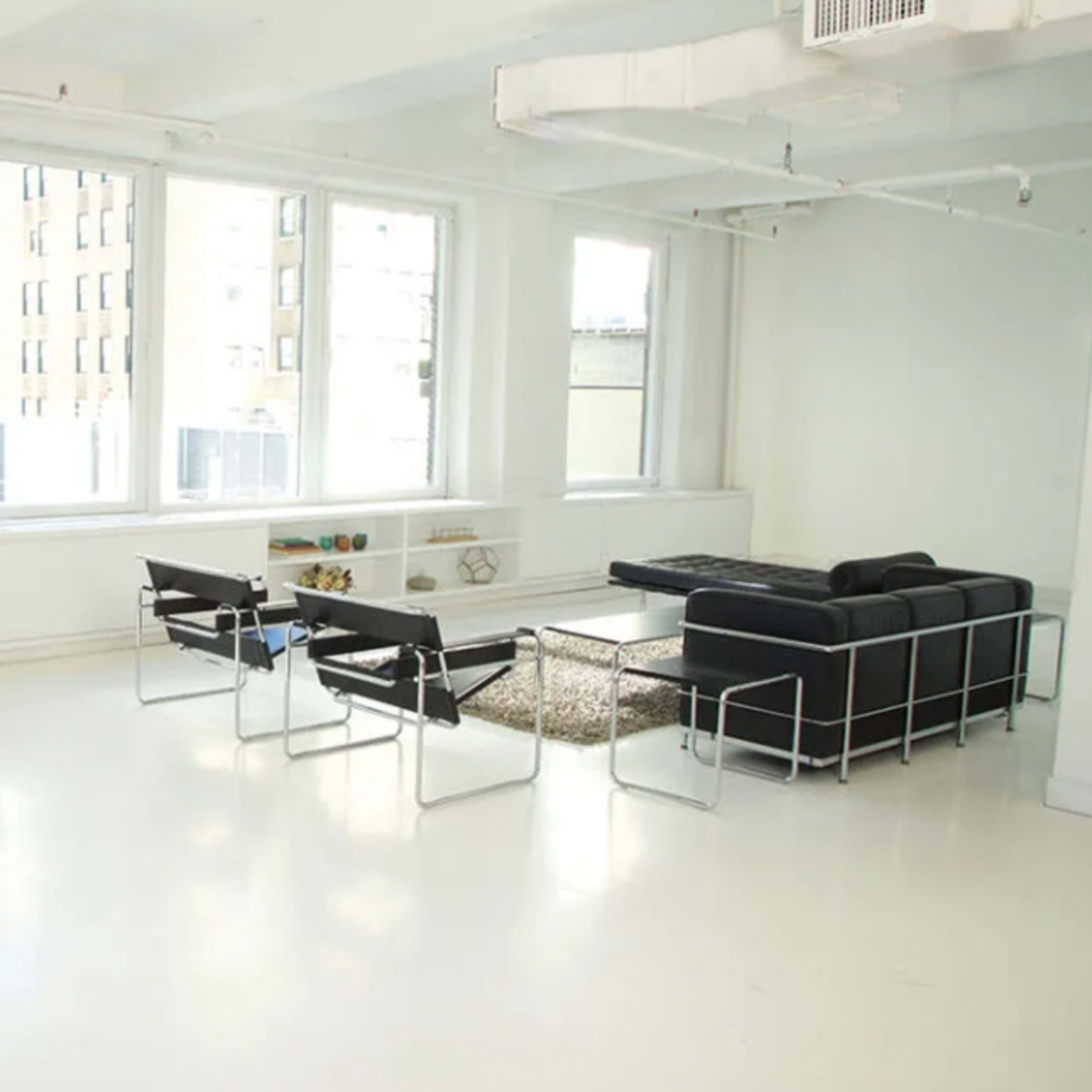 A minimalist living space features a black sofa, two chairs, a coffee table, and large windows providing natural light.