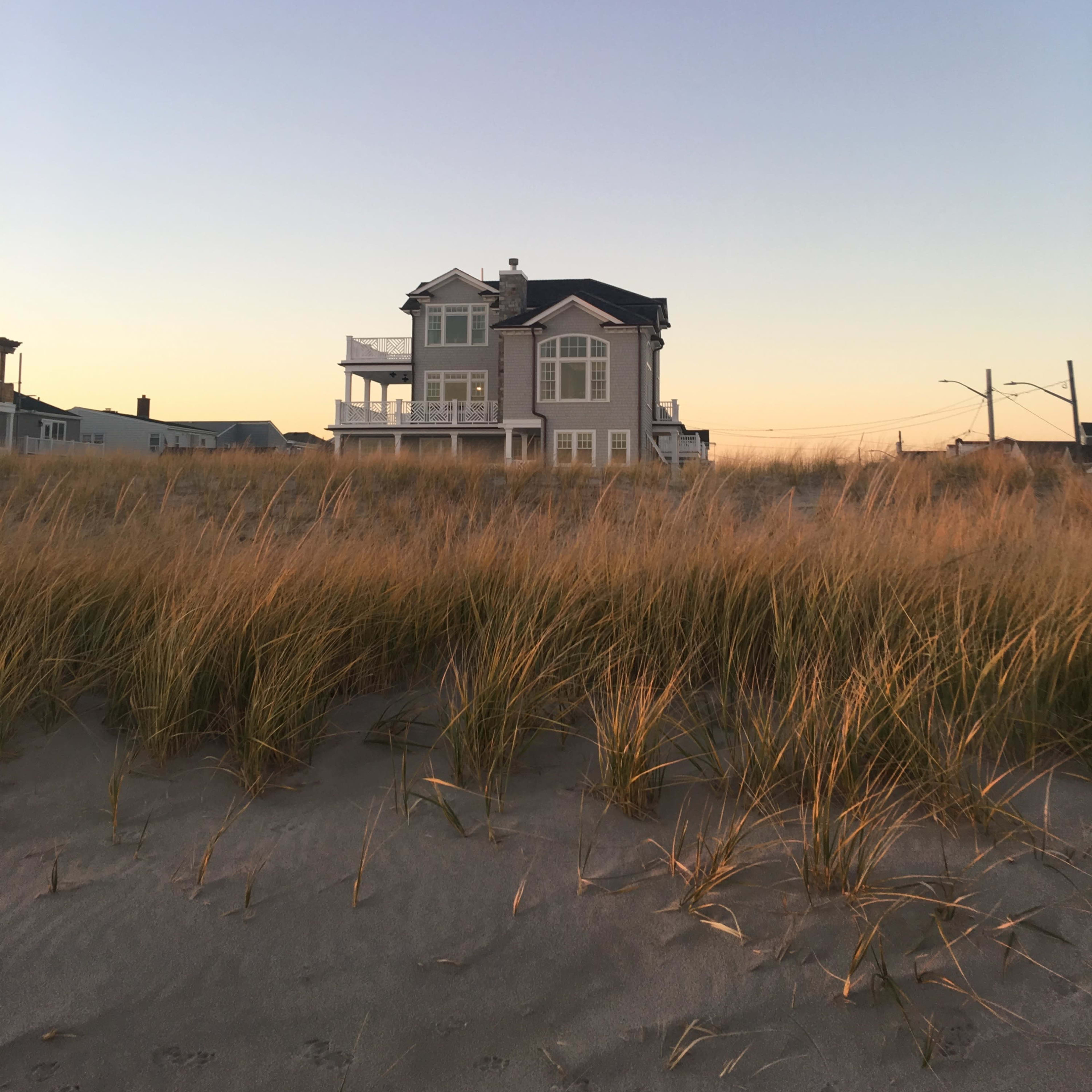 A beach house stands behind tall grasses at sunset, with clear skies in the background.