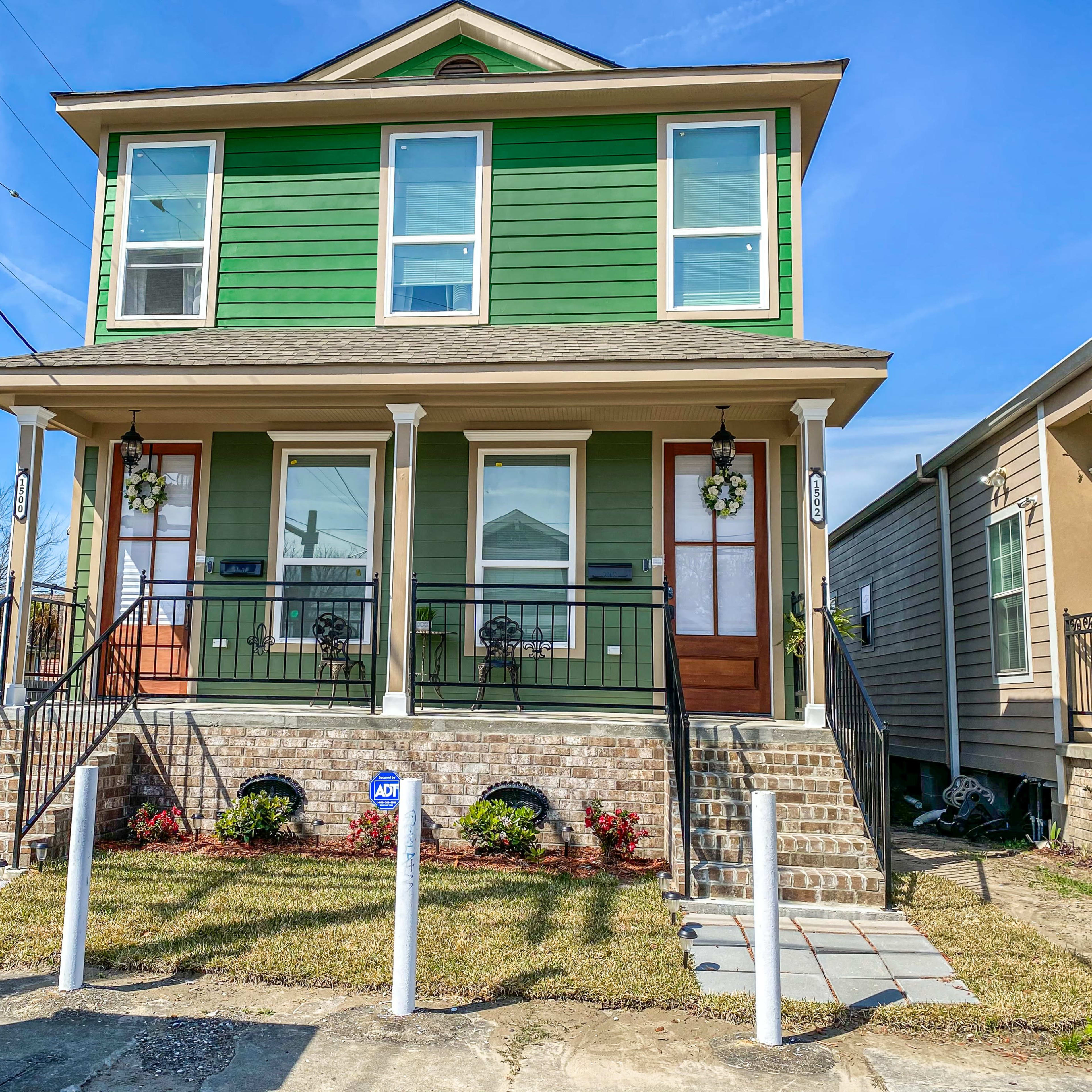 A two-story green duplex with a brick foundation features two entrance doors, each with a small porch and decorative plants.