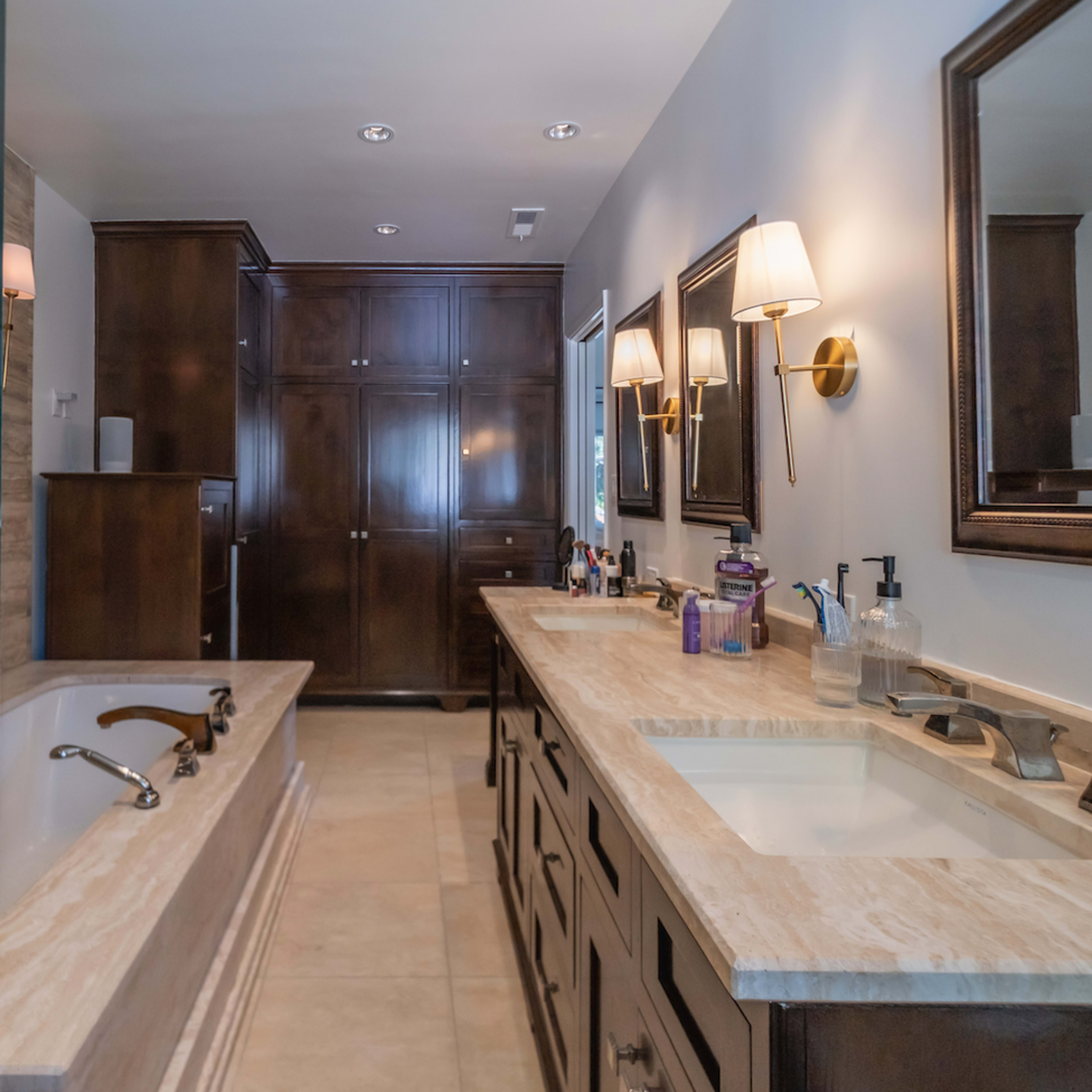A modern bathroom with a glass shower, a large bathtub, dual sinks with a marble countertop, and wooden cabinetry.