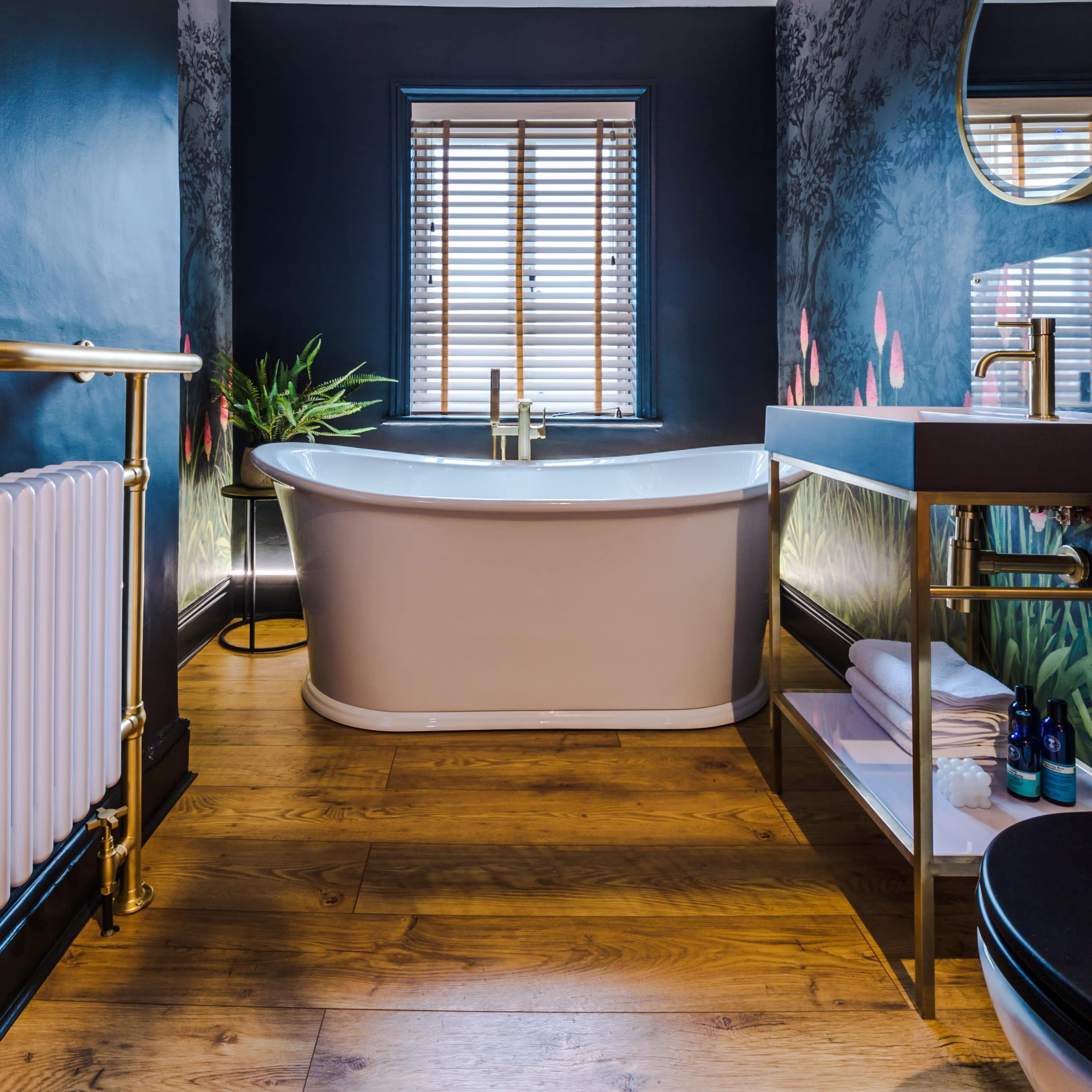 A modern bathroom featuring a freestanding bathtub, a round mirror, wooden flooring, and a window with blinds.