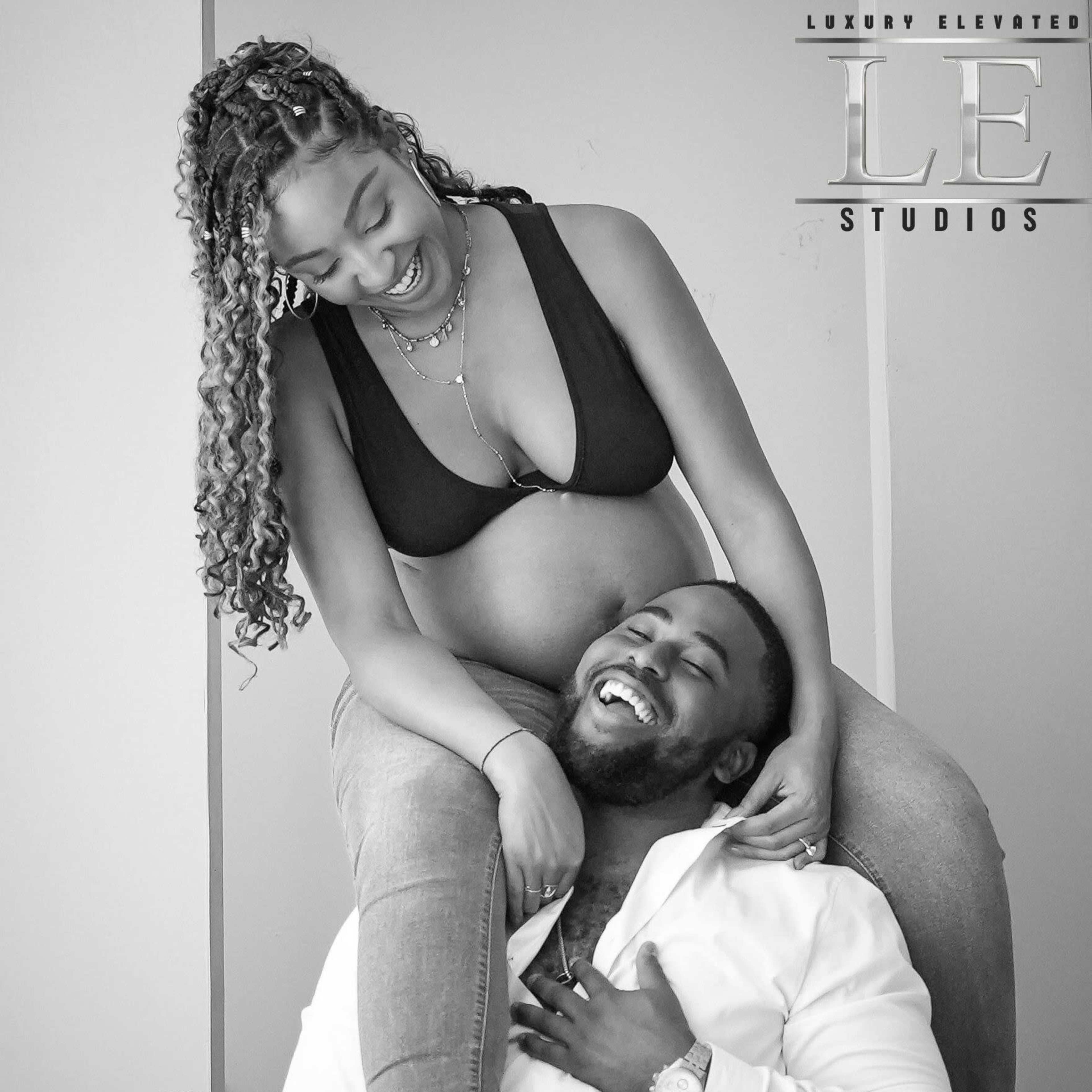 A pregnant woman smiles down at a man sitting on the floor while she playfully leans on him.