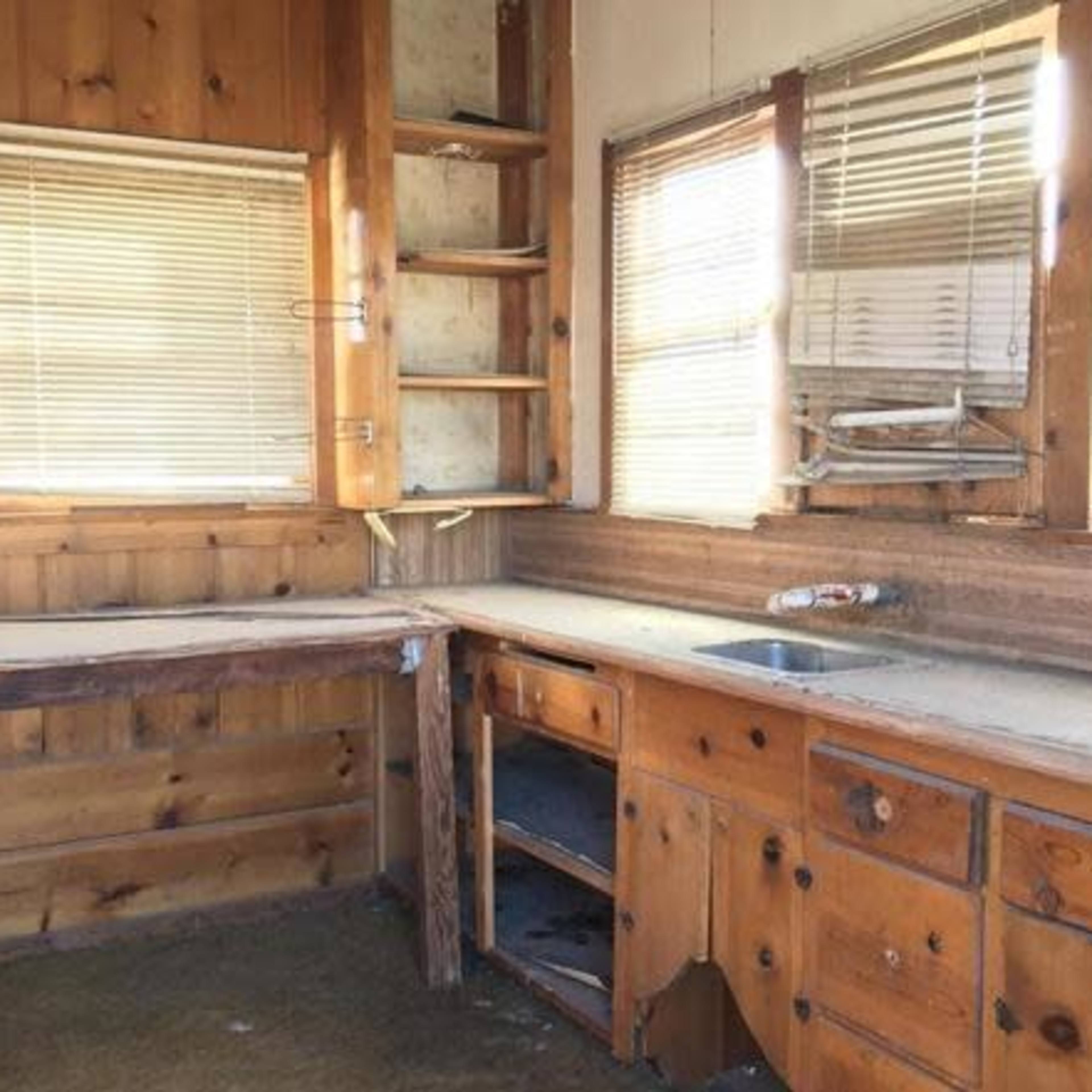 A wooden kitchen with a countertop, shelves, and a sink, featuring peeling paint and cluttered surfaces.