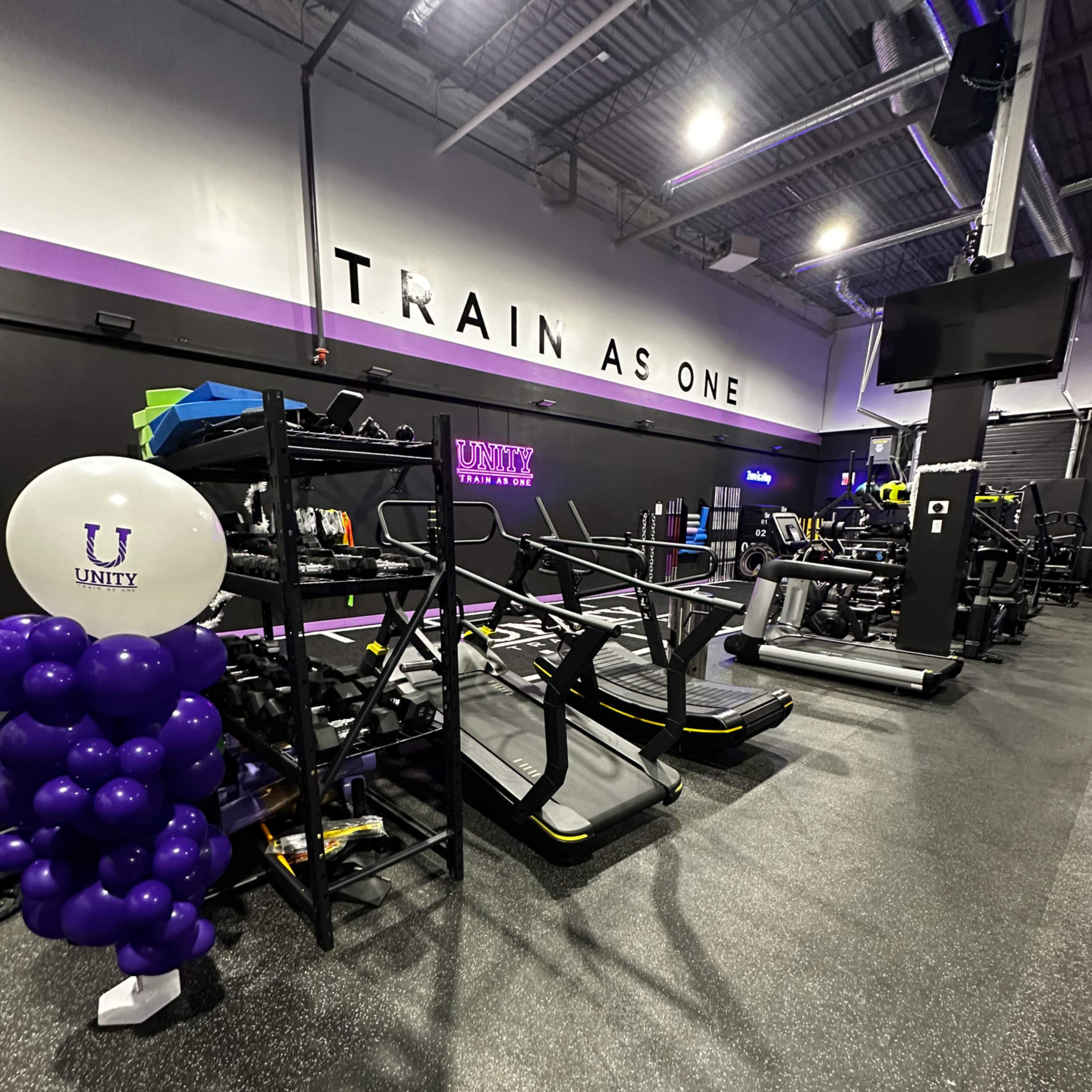 A gym interior with exercise equipment lined up along the wall, a balloon display, and the phrase "TRAIN AS ONE" prominently on the wall.