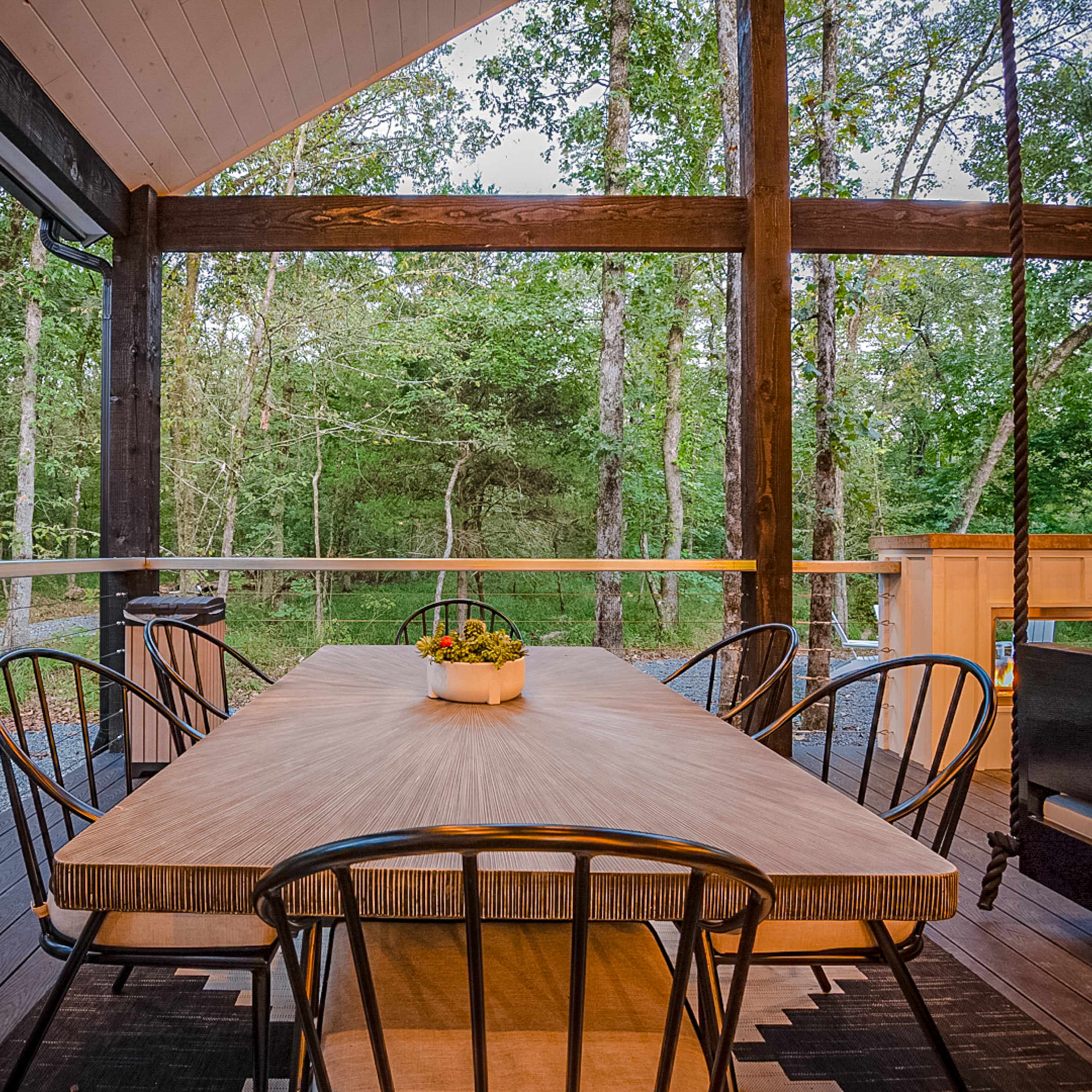 A rustic outdoor dining area features a wooden table surrounded by black chairs, with a view of a wooded area in the background.