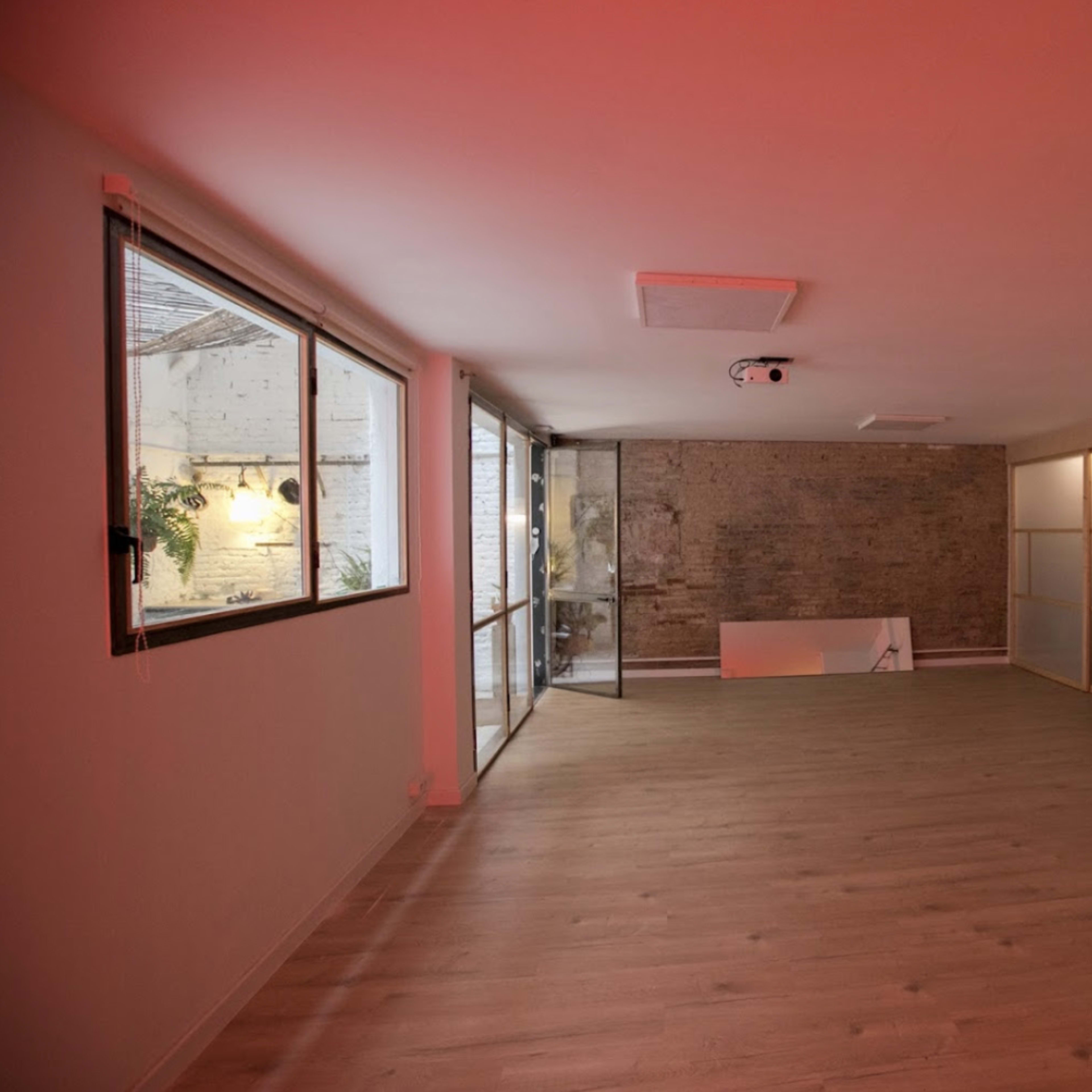 An empty room with wooden flooring, a brick wall, large windows, and minimal furnishings, illuminated by soft red lighting.