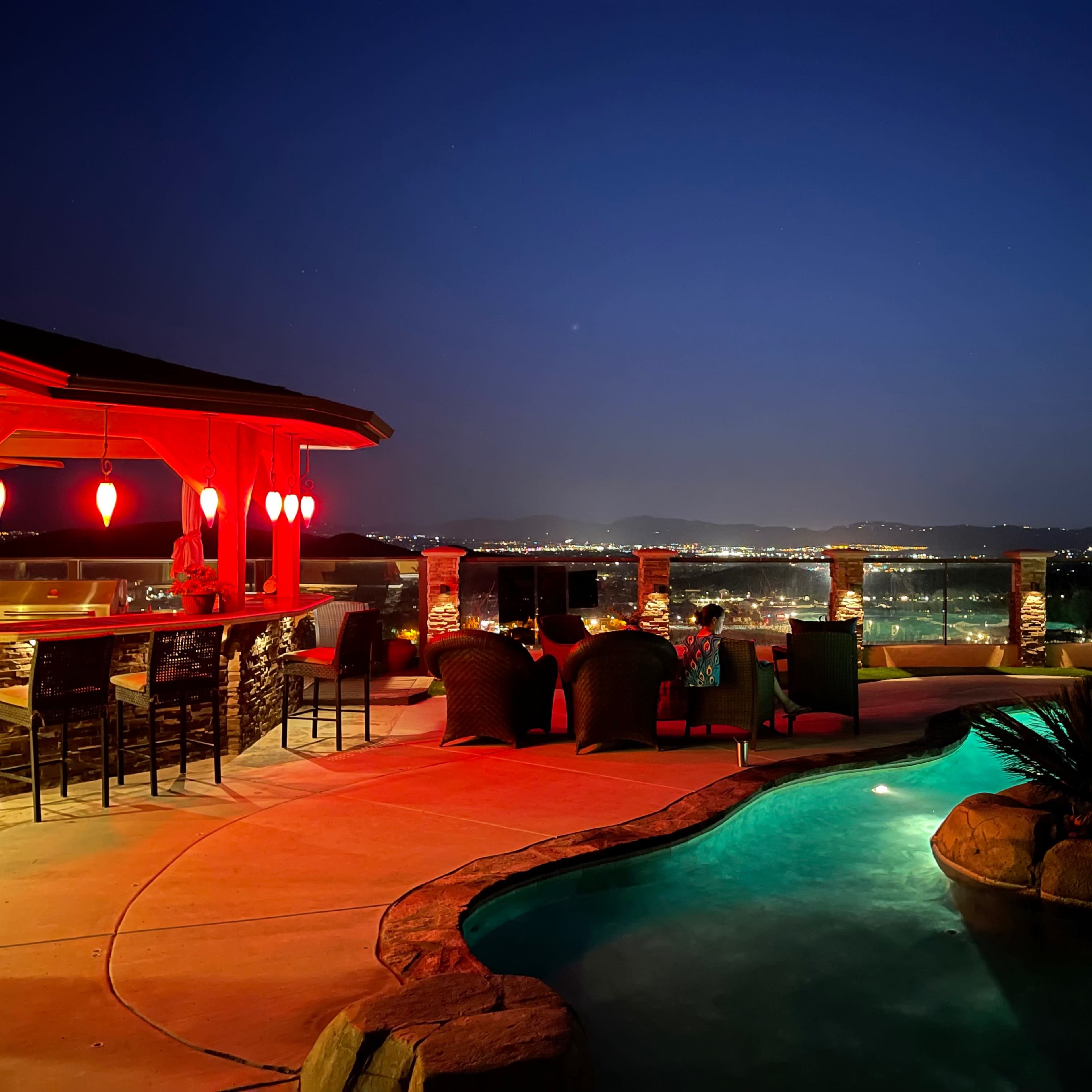 A luxurious backyard patio features a lit pool, a bar area, and panoramic views of the city at night.
