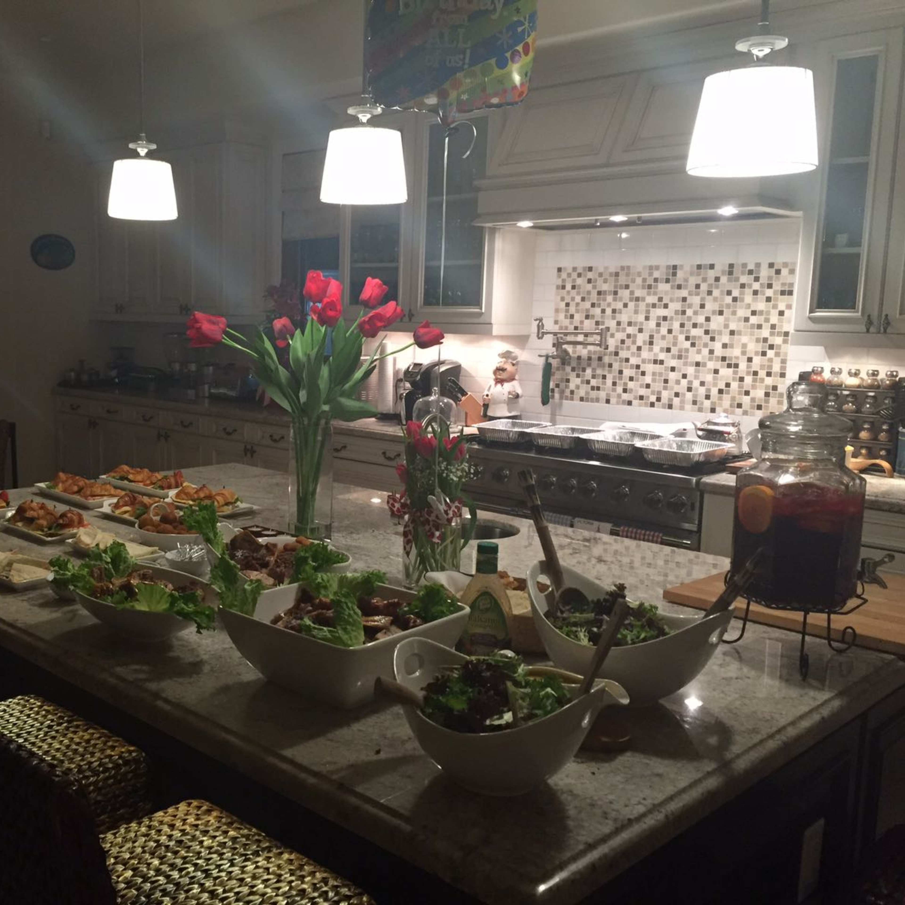 A well-lit kitchen features a large island with various dishes of food, fresh salads, and a vase of red tulips in the center.