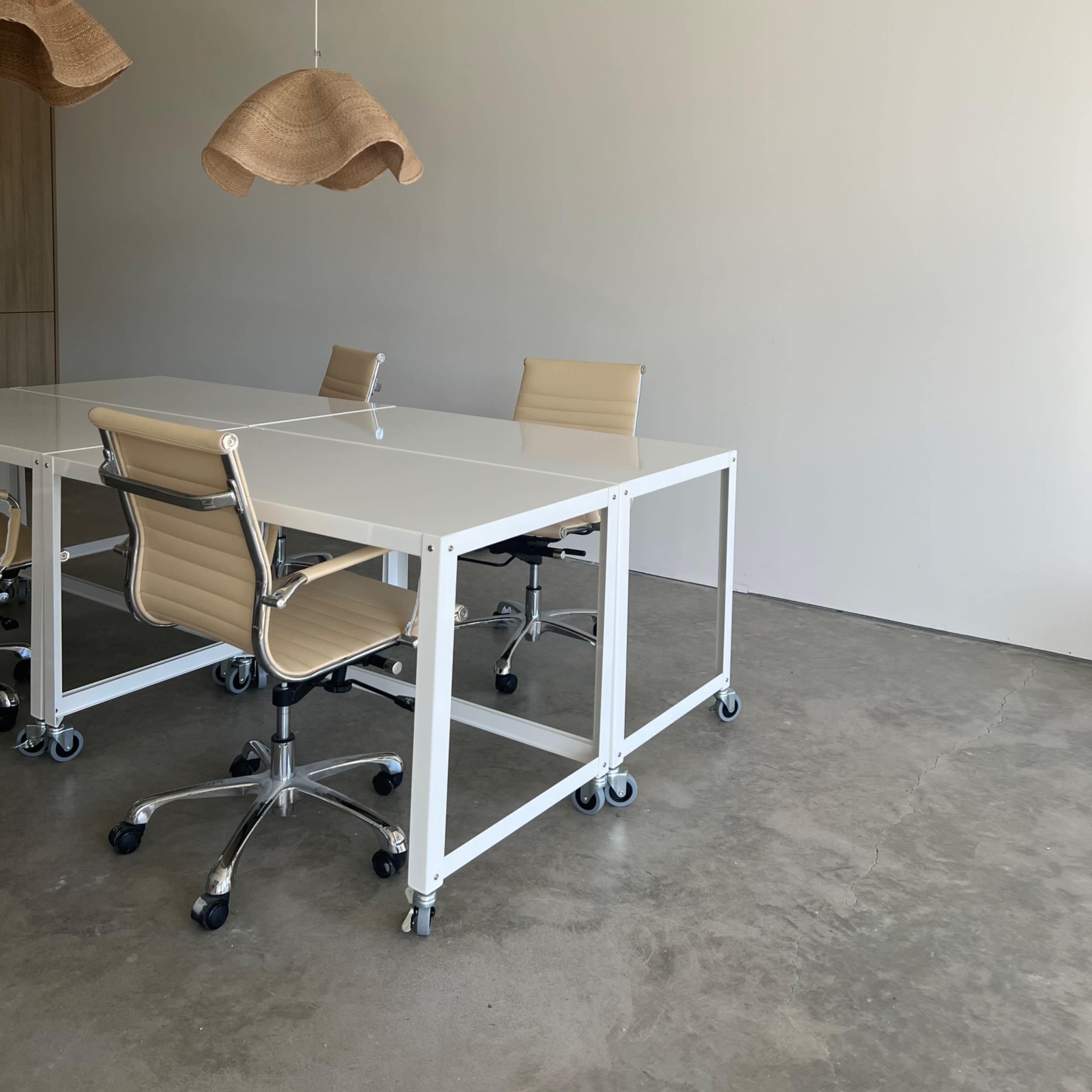 A minimalist conference room features a white table and beige rolling chairs beneath two woven pendant lights.