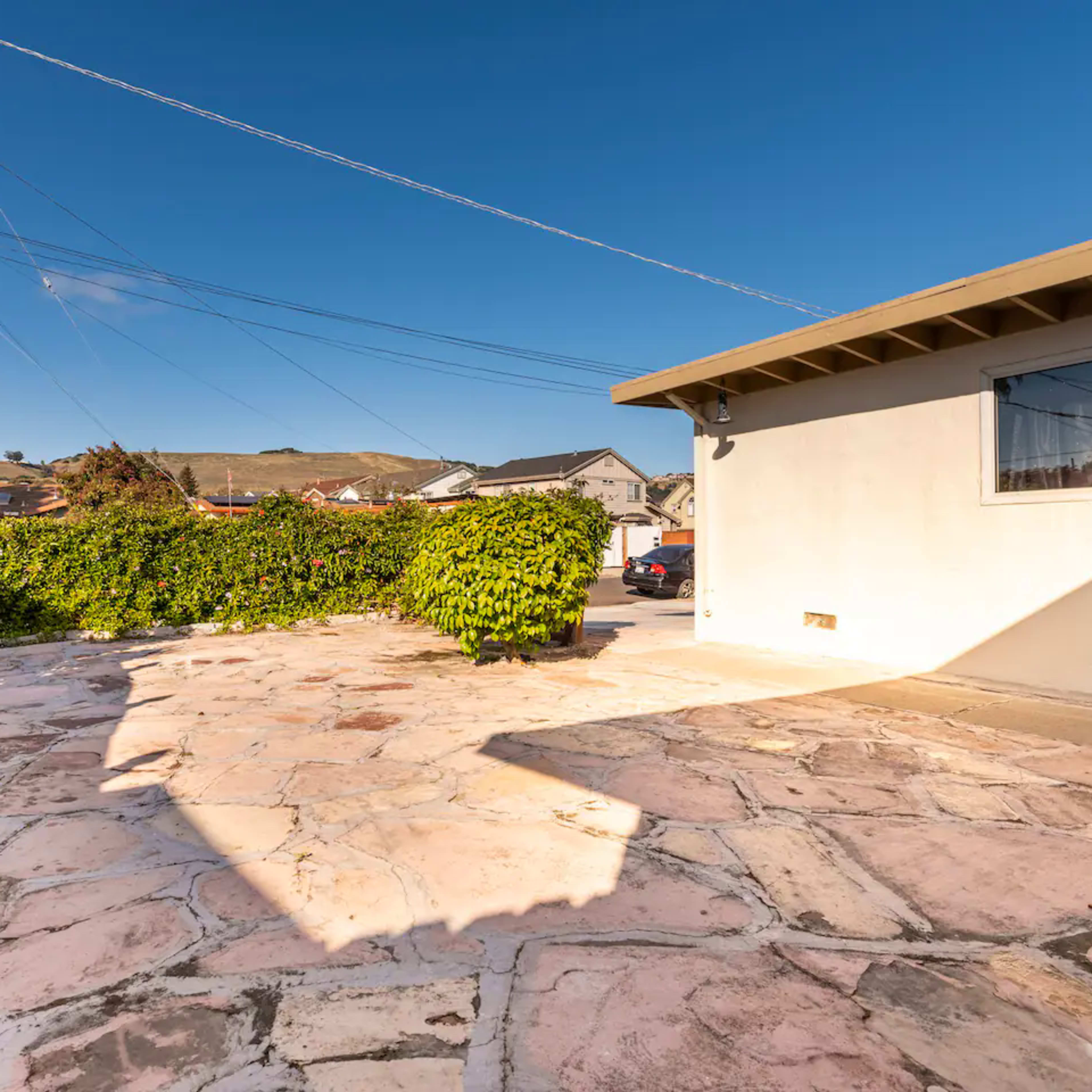 A stone patio with a shadow cast by a wall, surrounded by greenery and power lines in a residential area.