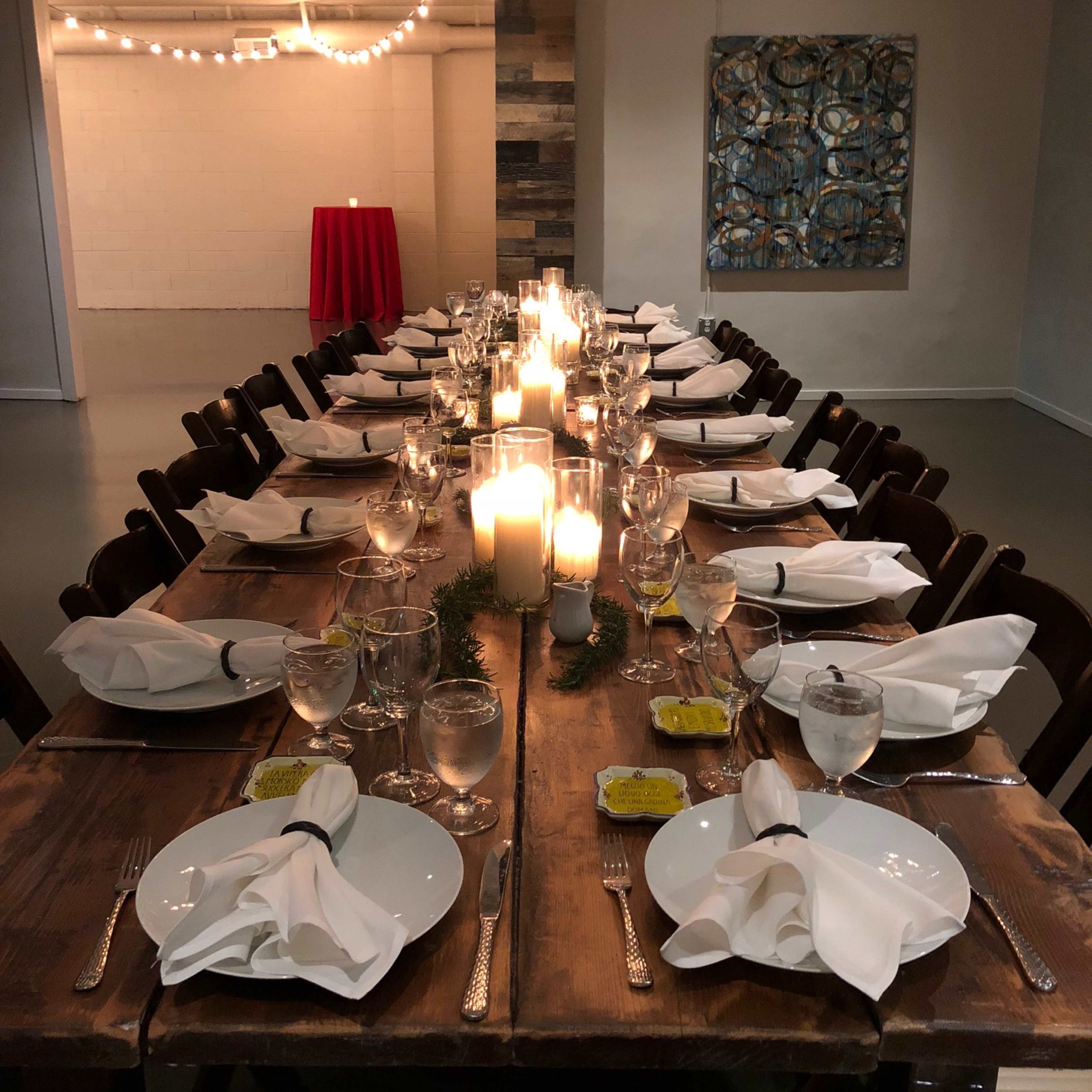 A long wooden dining table is set with white plates, folded napkins, water glasses, and lit candles, under decorative light fixtures and wall art.