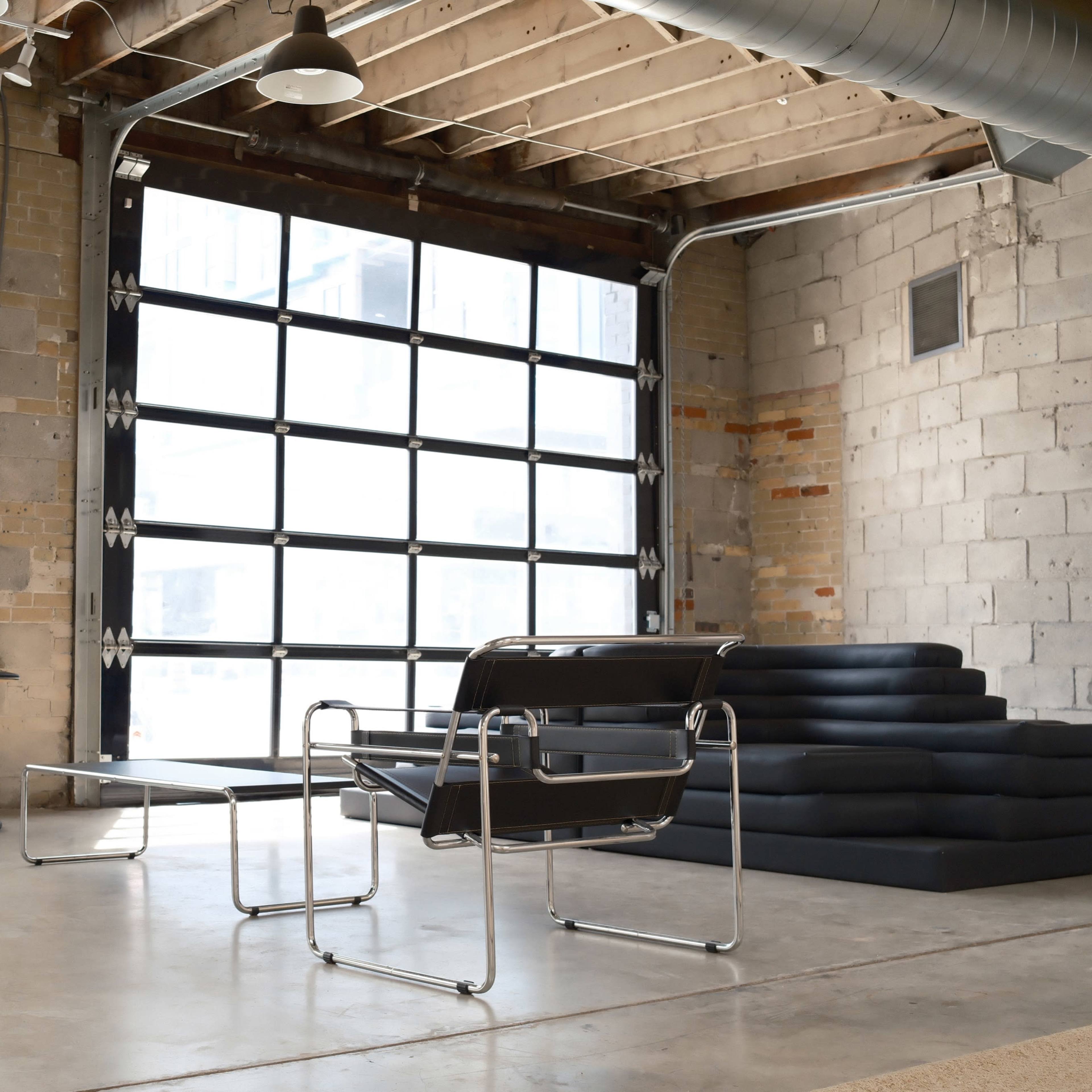 A modern, open room with a large garage door, a window with sheer curtains, two black chairs, and a stack of dark cushions on the floor.