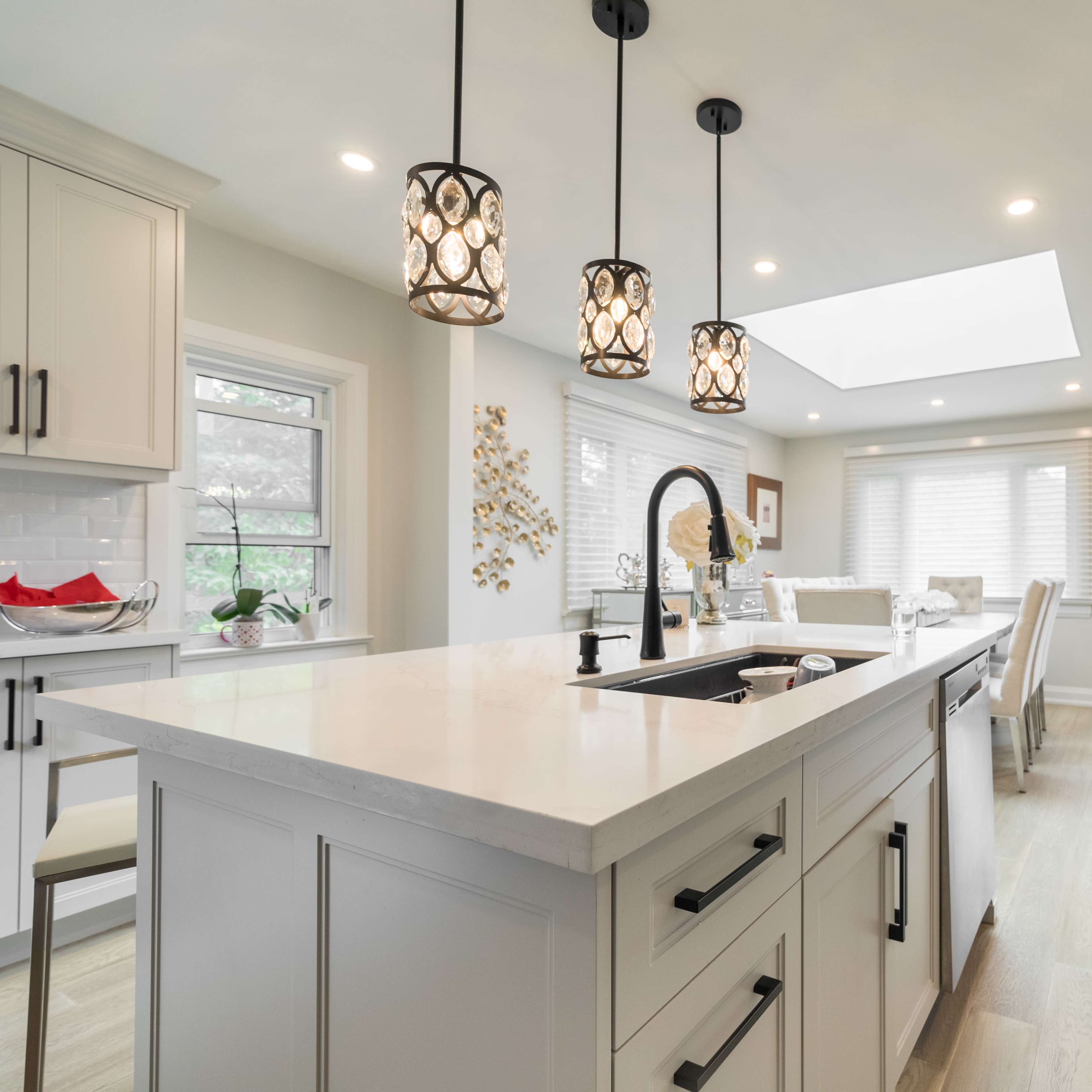 A modern kitchen with light-colored cabinetry, a large island with a sink, and pendant lights above, leading to a dining area with a skylight.