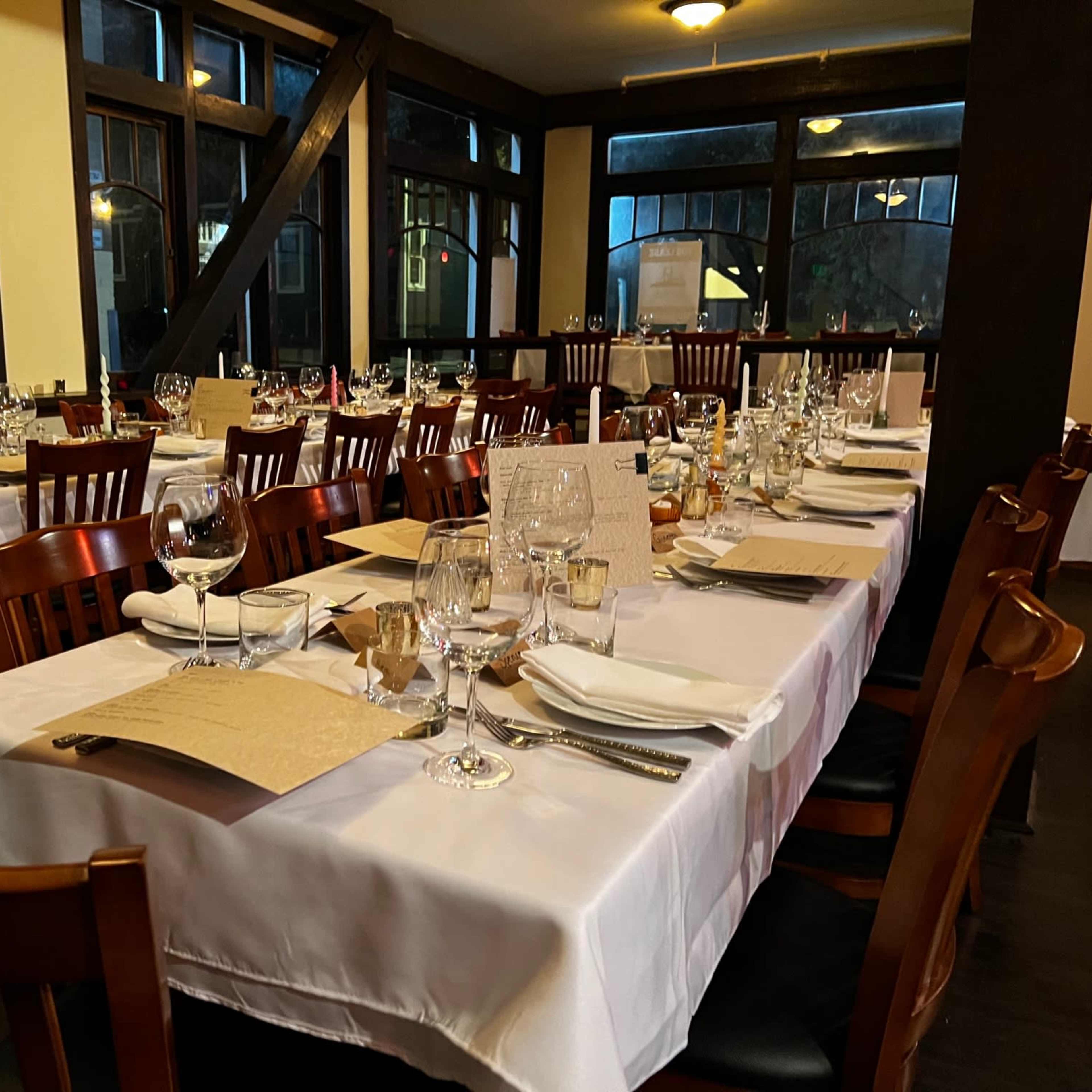 A row of neatly set tables with white tablecloths and tableware is arranged in a well-lit restaurant dining area.