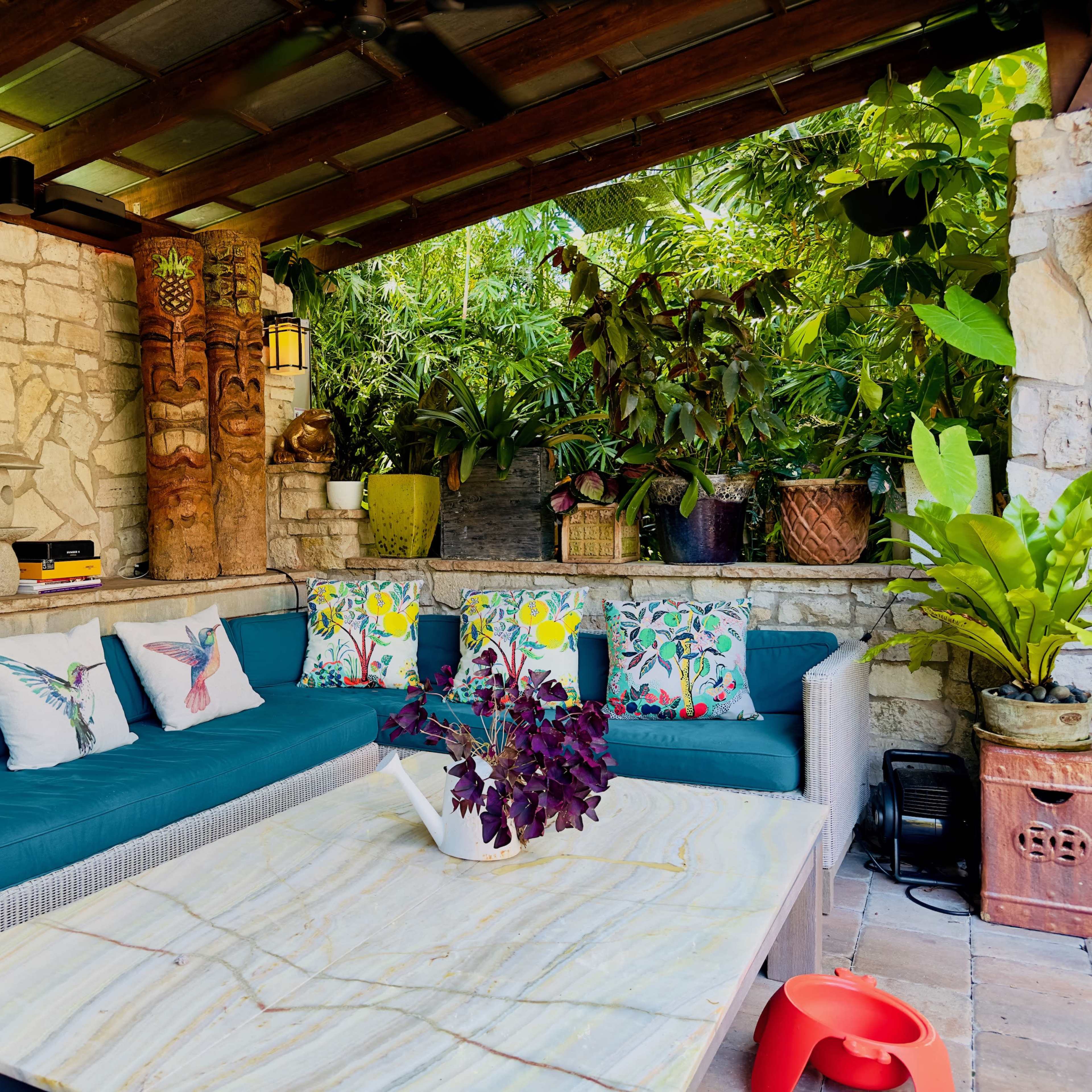 A patio area featuring a blue cushioned sectional sofa adorned with decorative pillows and a marble coffee table, surrounded by lush greenery and various potted plants.