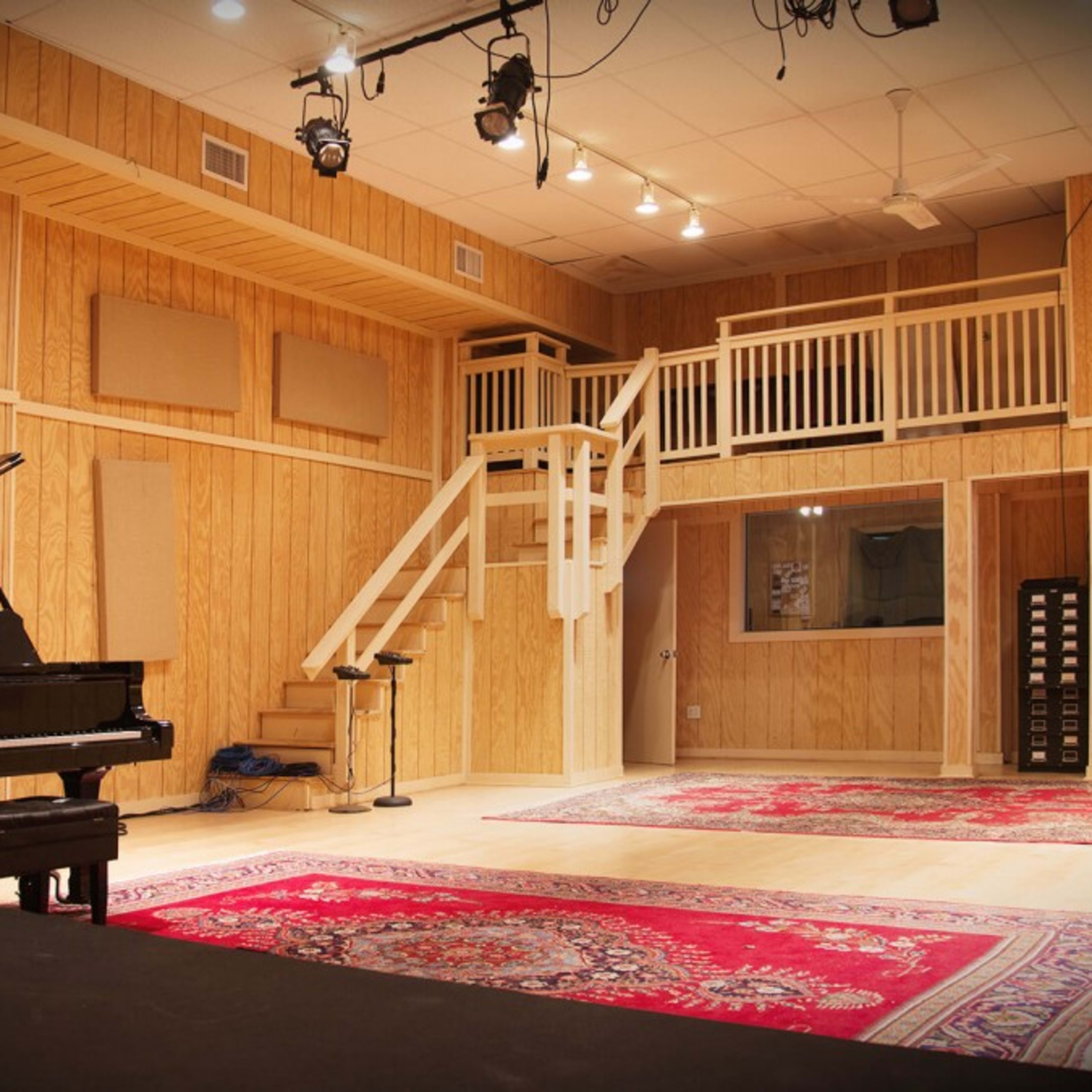 A music studio featuring a grand piano, wooden walls, a staircase, and carpeted areas with large rugs.