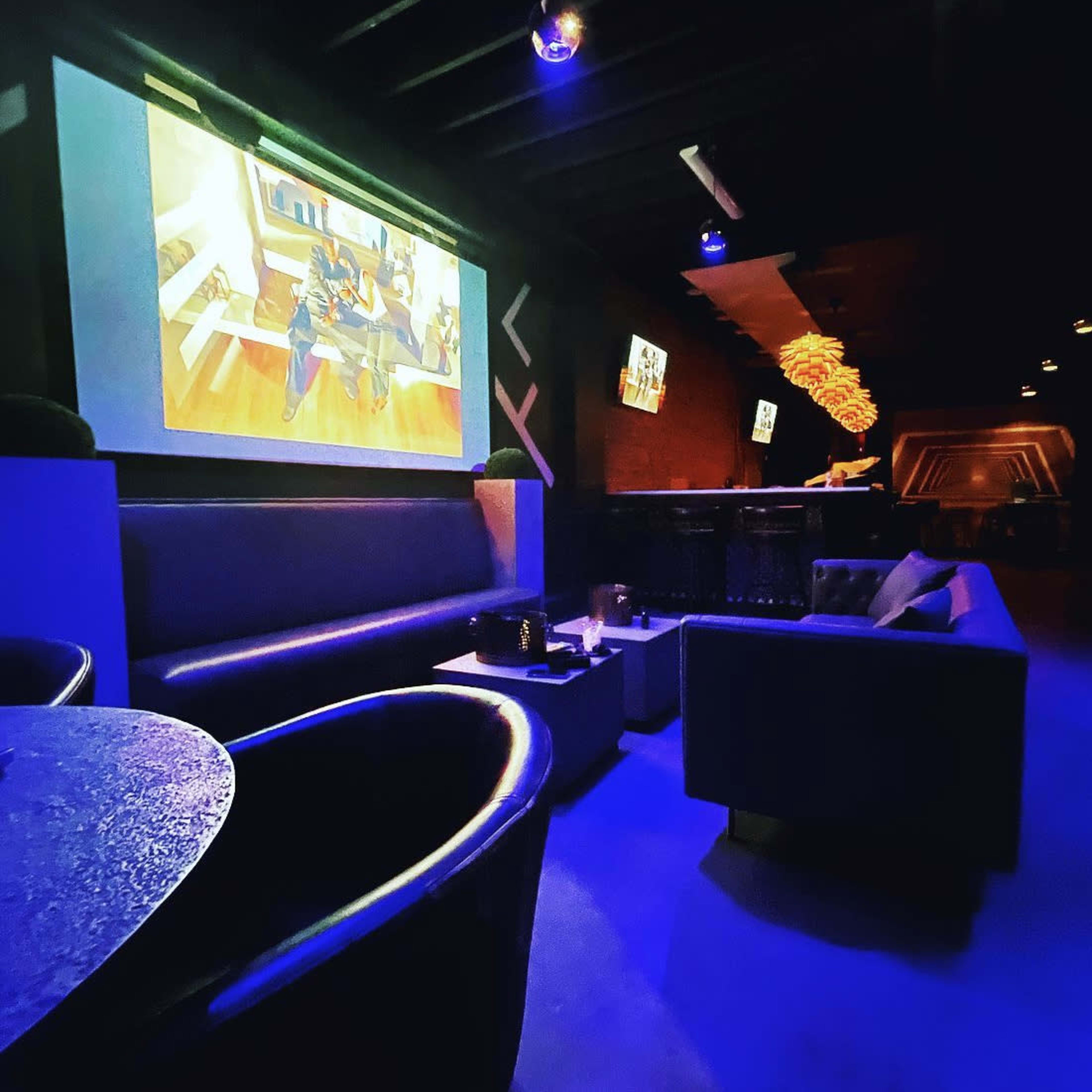 A dimly lit lounge area with dark furniture and a large screen displaying an animated scene on the wall.